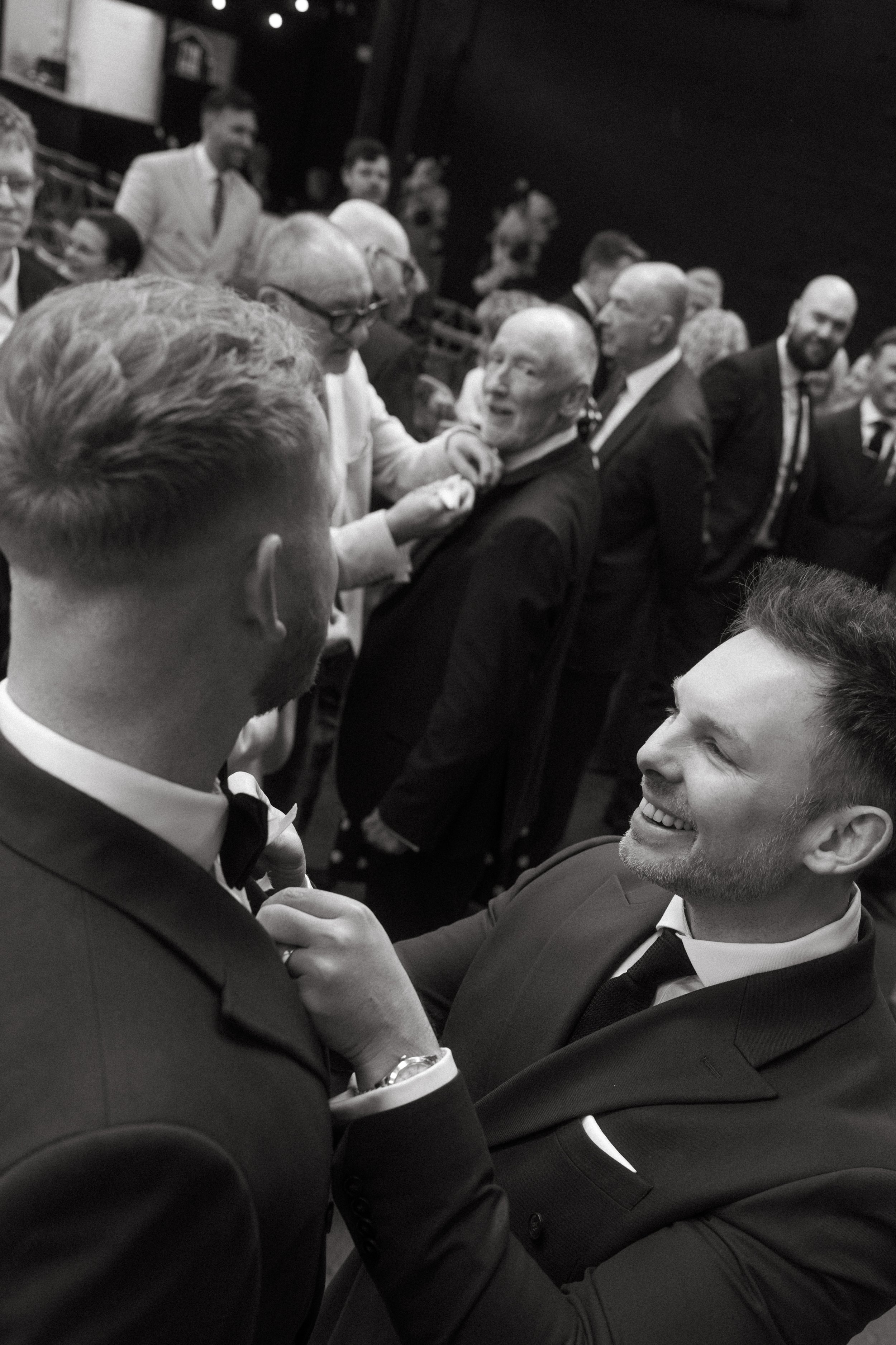 A formal event with people dressed in tuxedos, socializing and smiling. Two men in the foreground are shaking hands and appear to be enjoying a conversation.