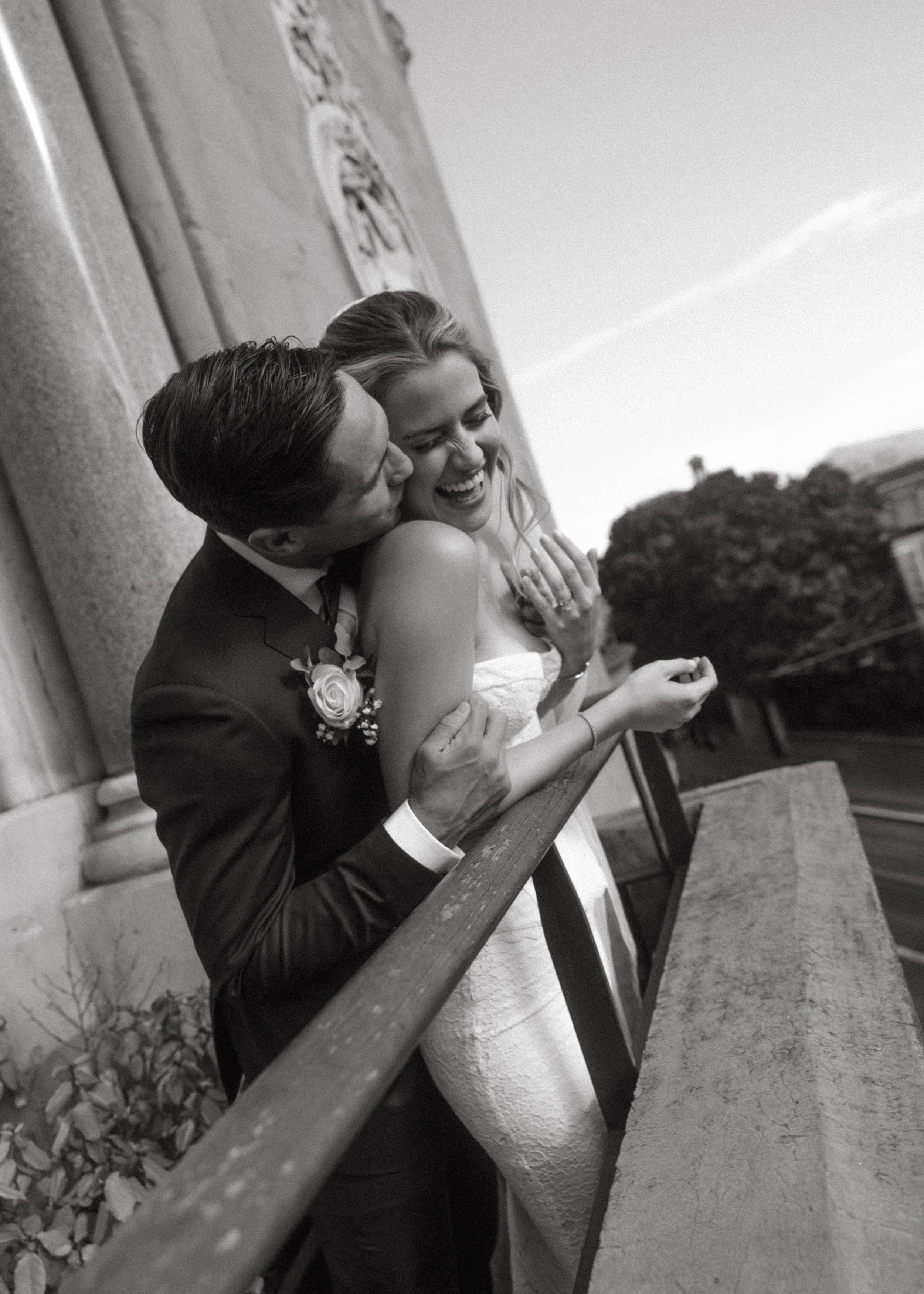 A black and white photo of a joyful bride and groom embracing outdoors near a stone wall, with the groom kissing the bride on the cheek and the bride smiling happily, wearing a wedding dress and a ring.