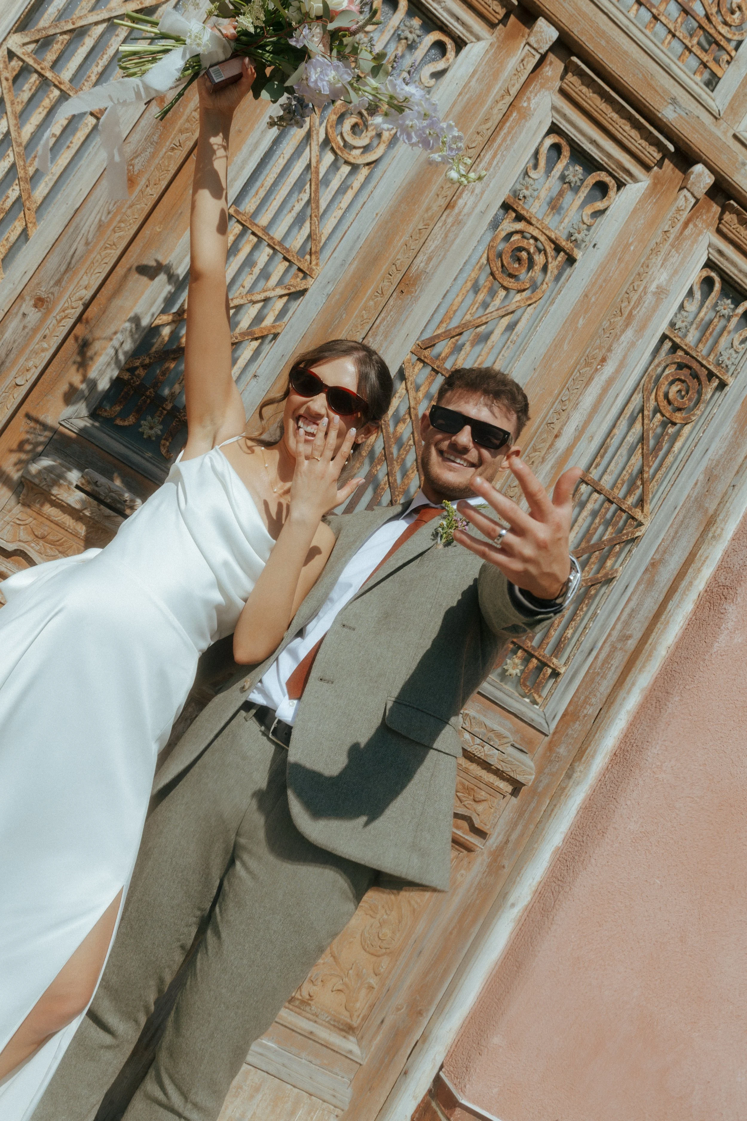 A happy couple taking a selfie together outdoors in front of an ornate wooden wall. The woman is wearing a white dress, sunglasses, and holding a bouquet of flowers overhead. The man is wearing a grey suit and sunglasses, showing a wedding ring and a
