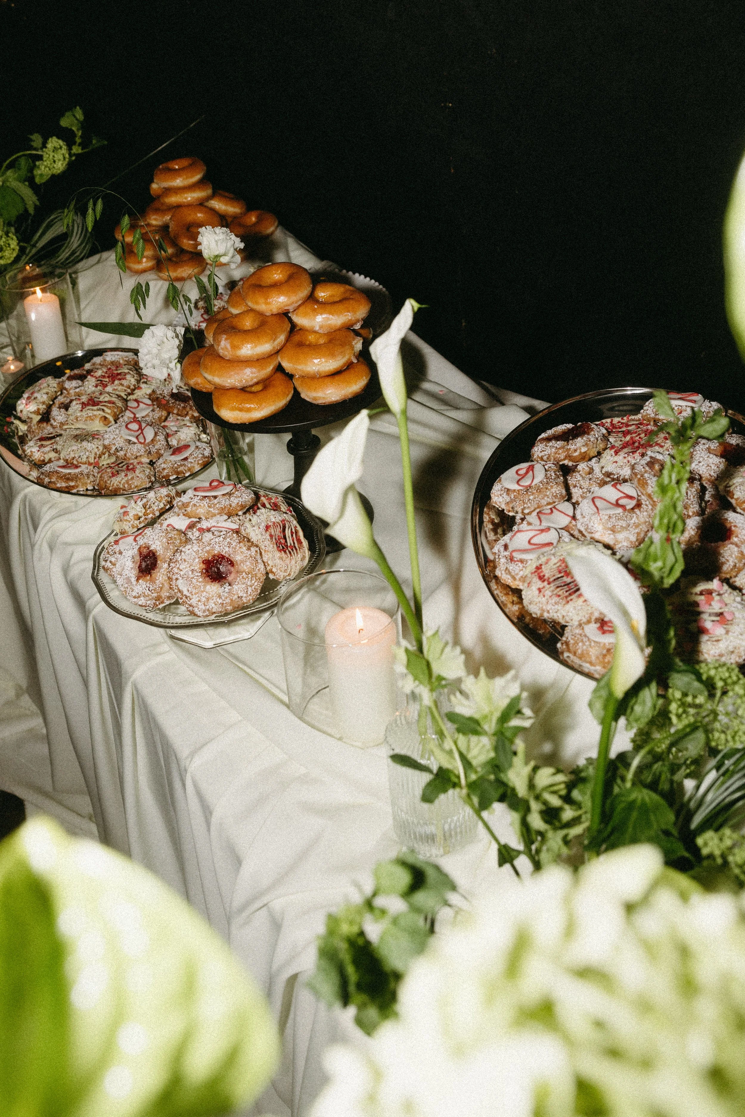 A dessert table with various cookies and donuts, decorated with white flowers and candles, draped with a white tablecloth and set against a black background.