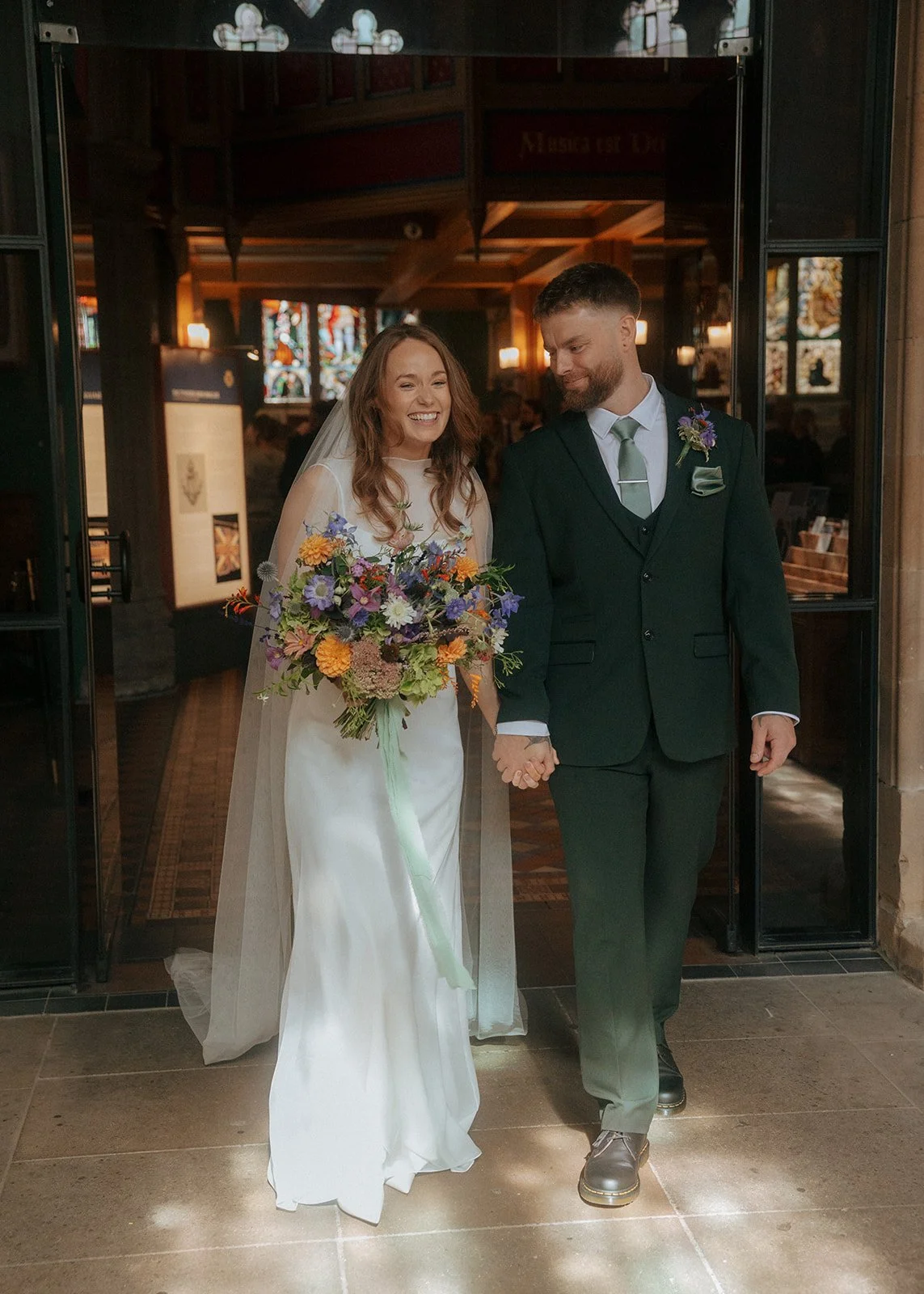 A bride and groom walking hand in hand at their wedding ceremony inside a church. The bride is holding a colorful bouquet of flowers and is smiling, while the groom is looking at her with a happy expression.