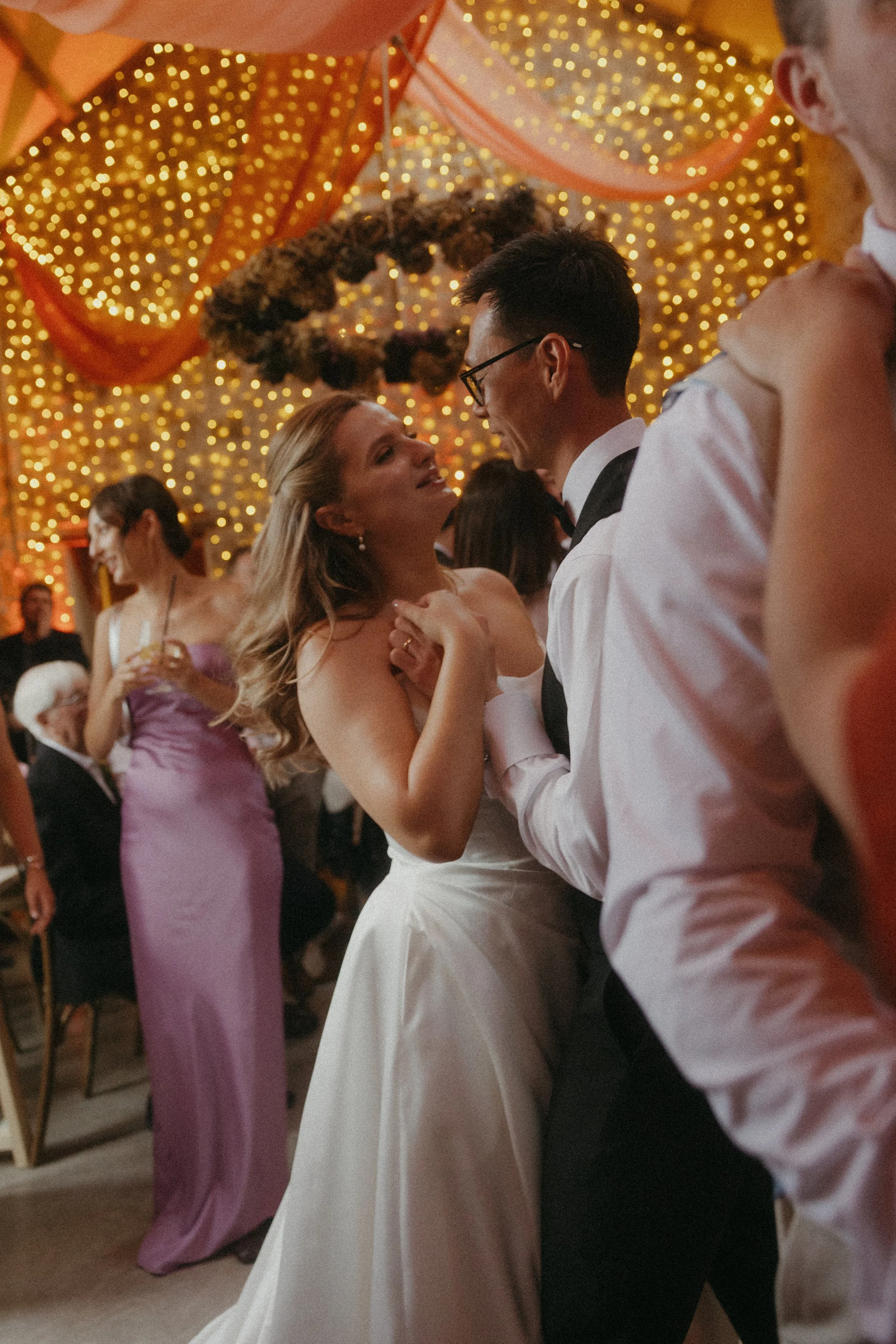 A bride and groom are dancing closely at their wedding reception, surrounded by guests, with decorative lights overhead.