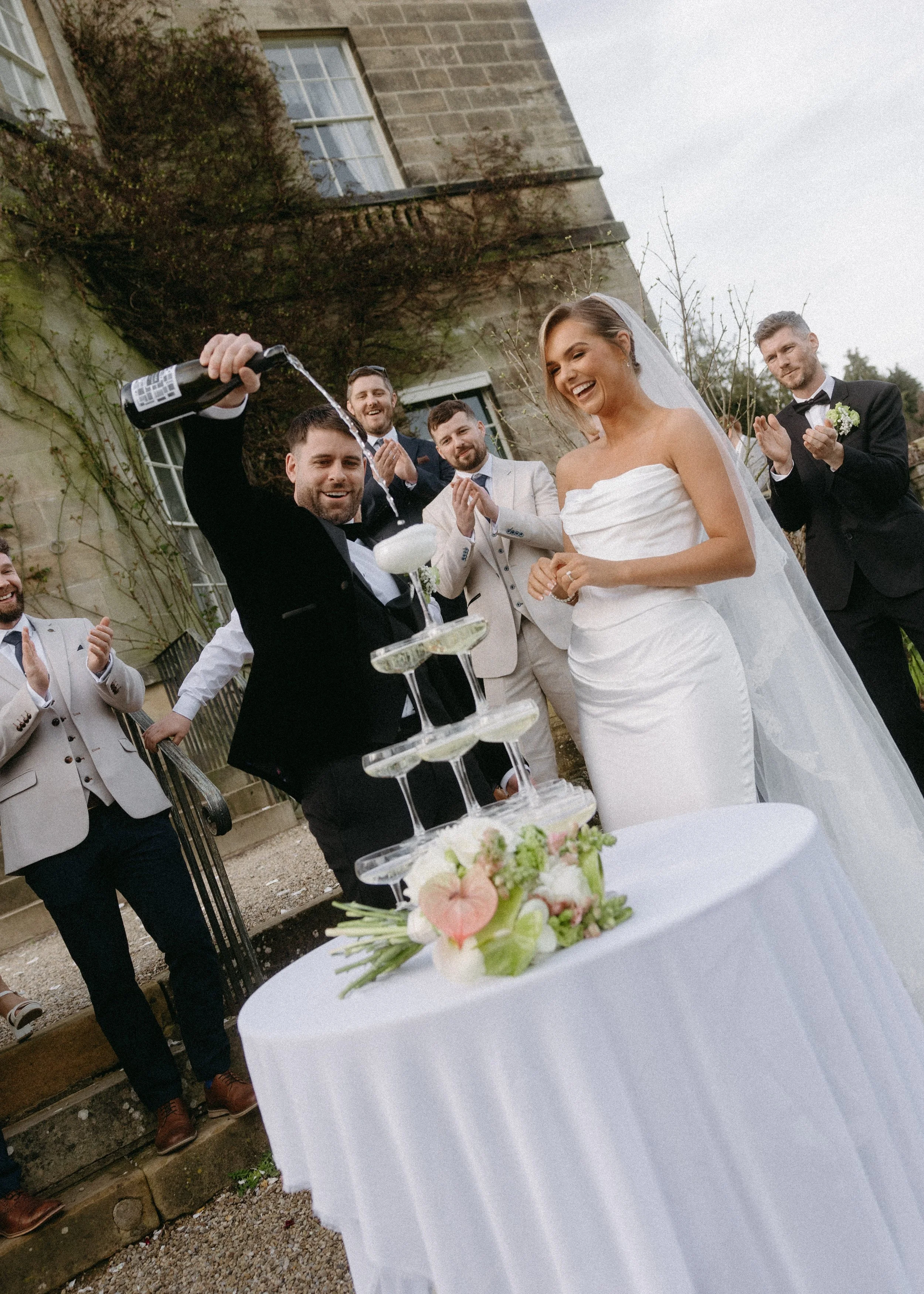 A wedding celebration with a bride and groom pouring champagne into a tower of glasses, while friends claps and cheers outdoors near a stone building with ivy.