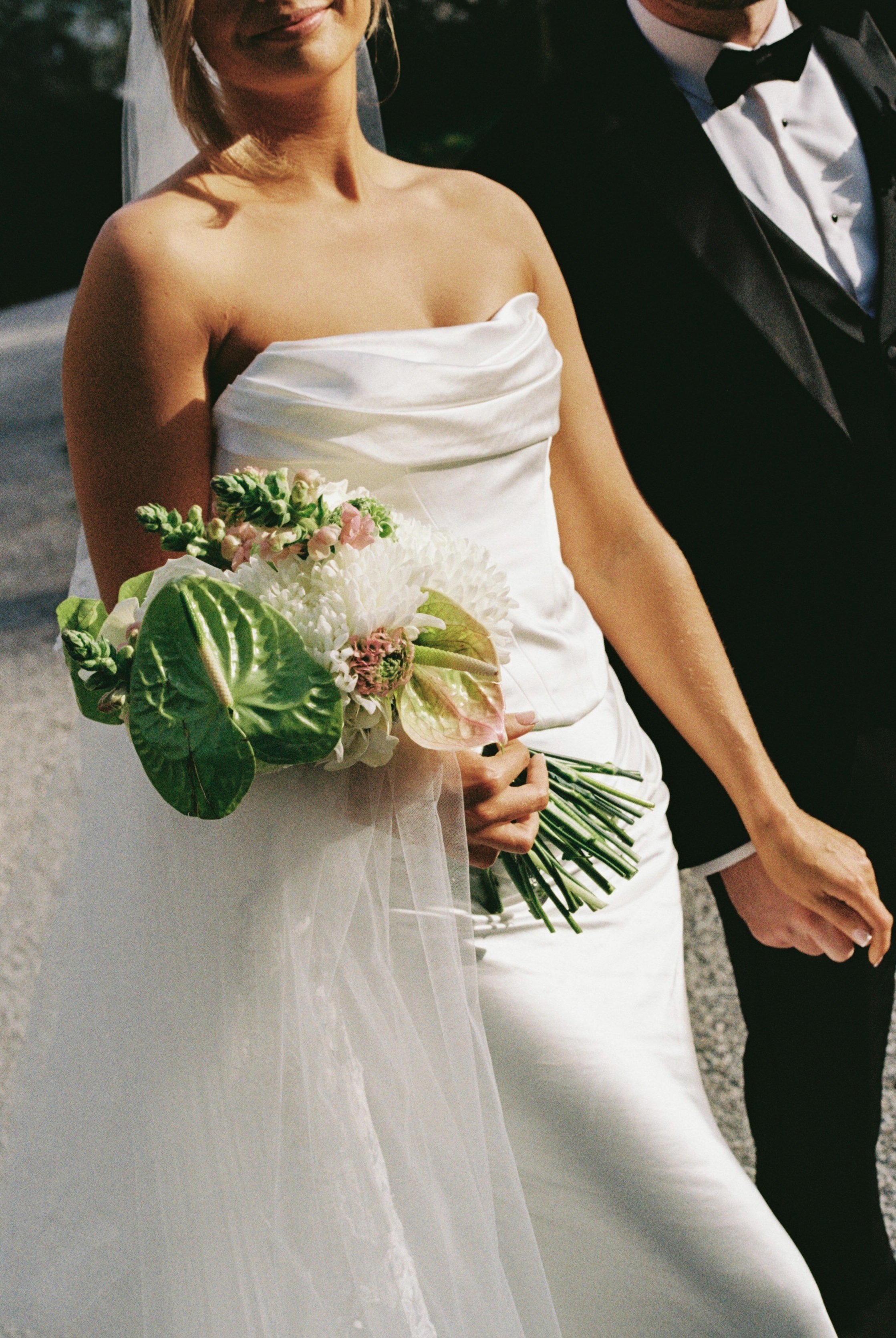 Close-up of a bride holding a bouquet of flowers, wearing a strapless white wedding gown, standing next to a groom in a black tuxedo with a bow tie.