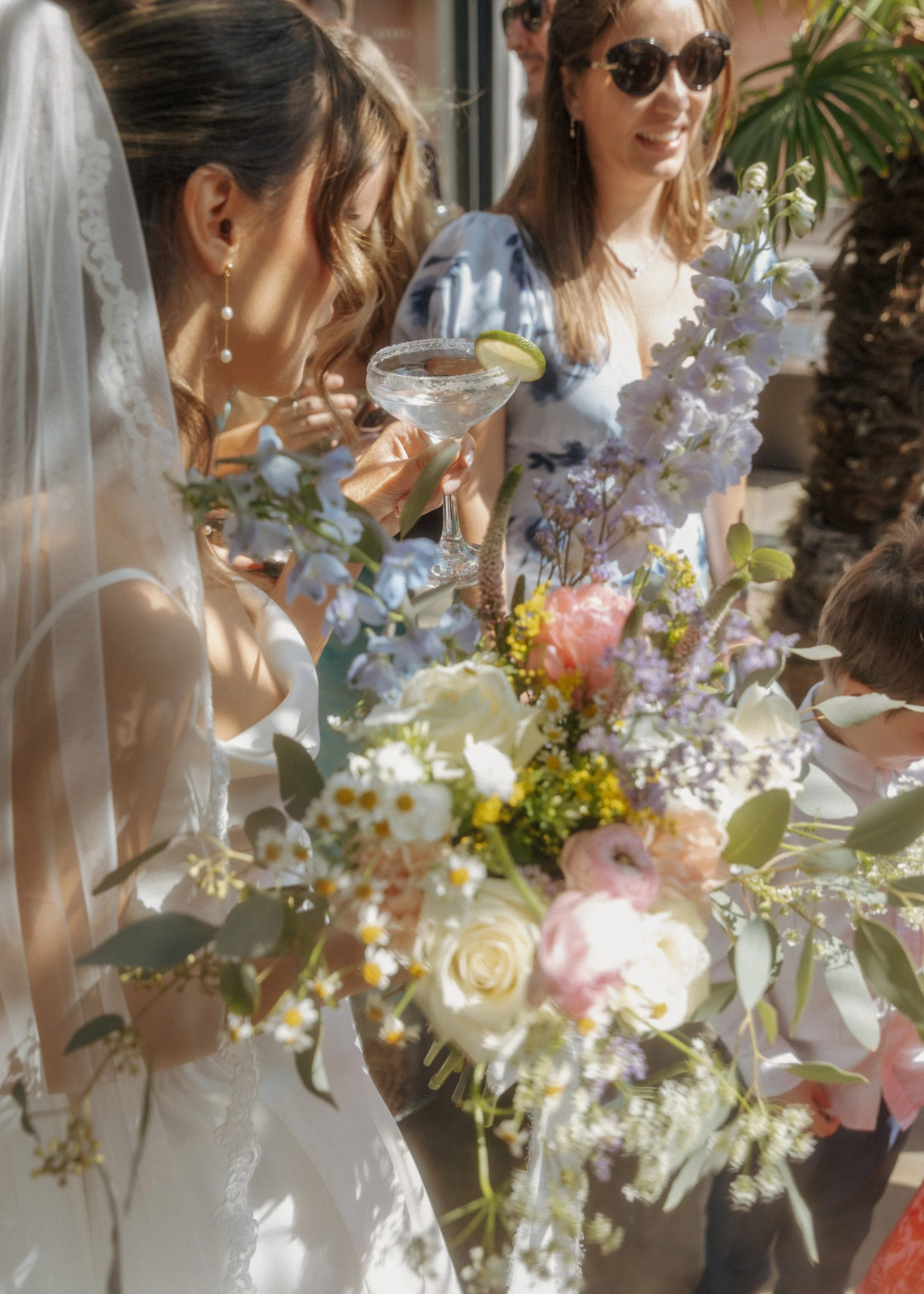 Women celebrating at a wedding reception, one holding a bouquet of flowers, another holding a cocktail glass with a lime wedge.