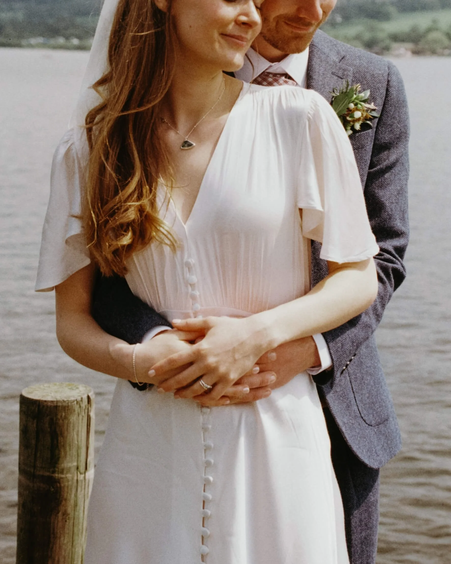 A couple dressed in wedding attire standing near a body of water, with the man standing behind the woman and holding her hands.