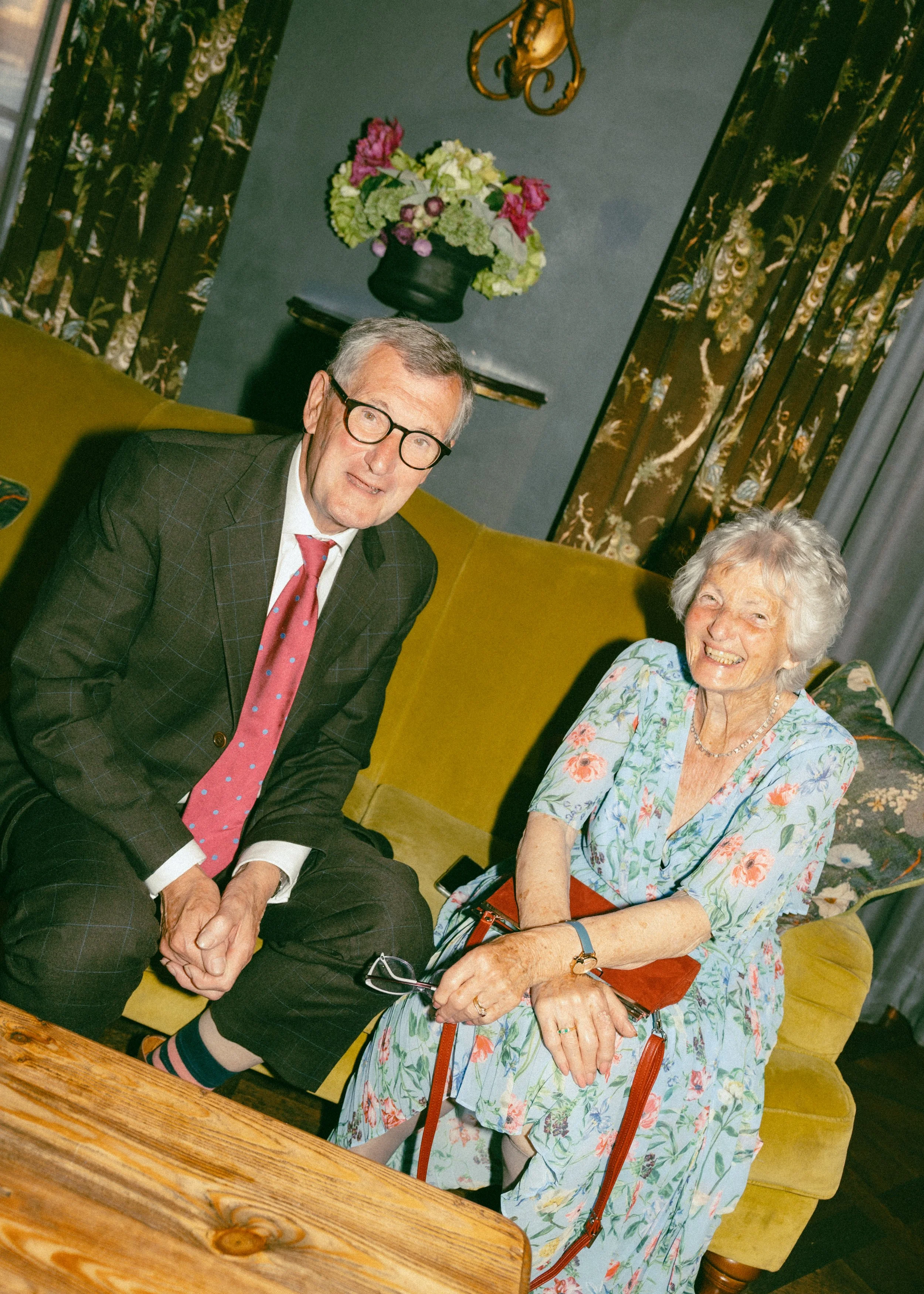 An elderly woman with gray hair wearing a floral dress is sitting on a yellow couch smiling. A man in a suit, glasses, and a red tie with blue polka dots is sitting next to her, smiling. There is a wooden table in front of them with eyeglasses placed