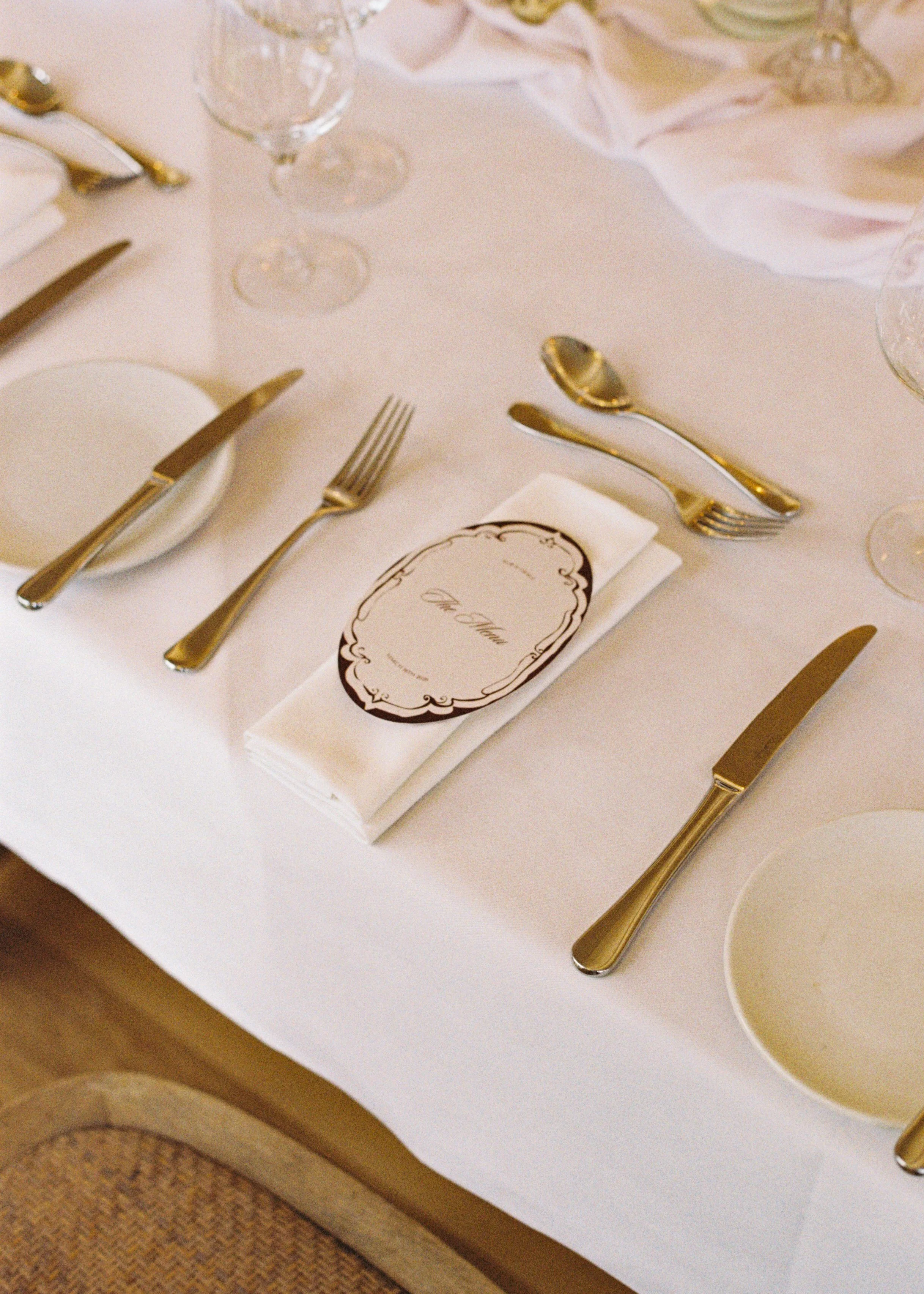 Elegant table setting with gold utensils, wine glasses, and a cloth napkin with a printed place card on a white tablecloth.