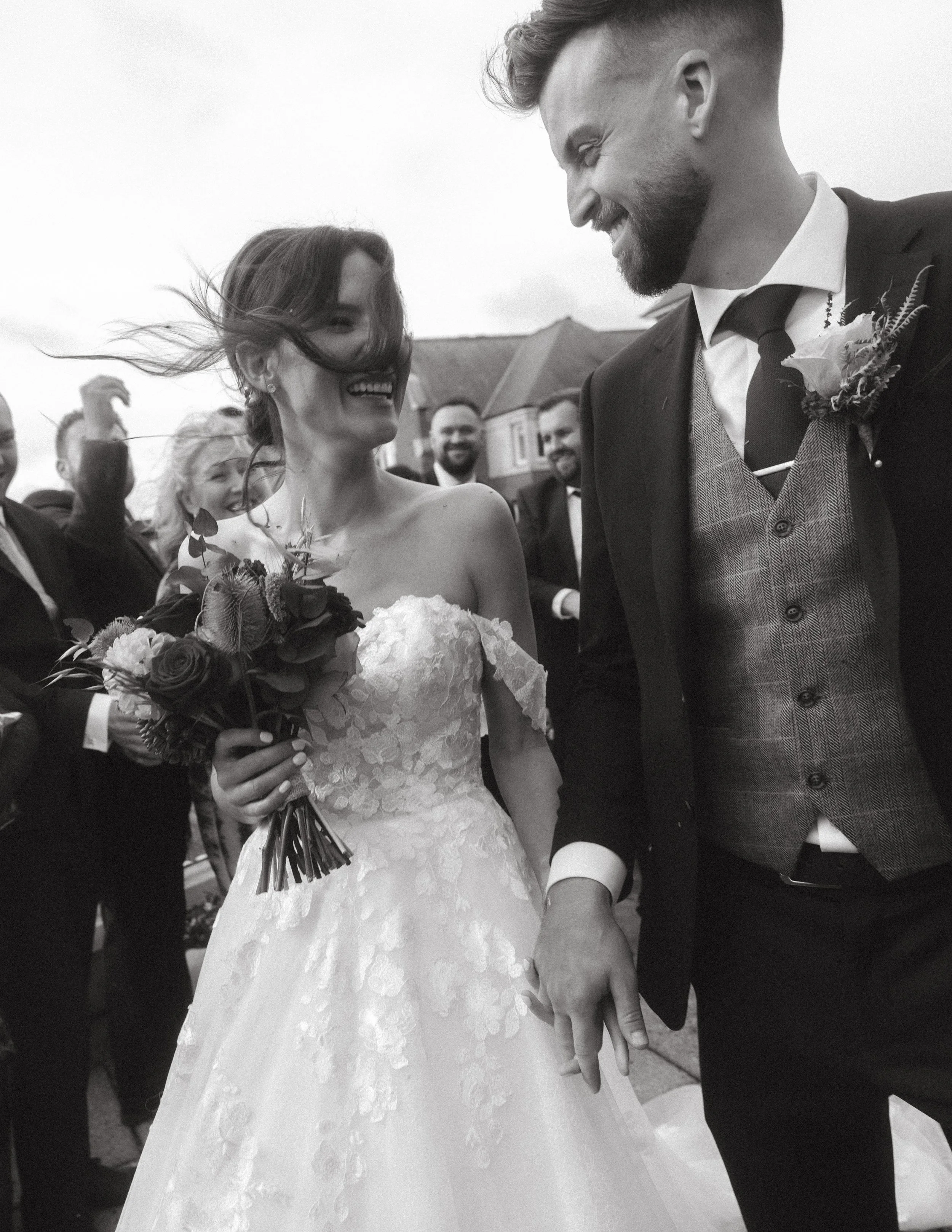 A smiling bride holding a bouquet of flowers, standing next to a man in a suit during a wedding celebration.