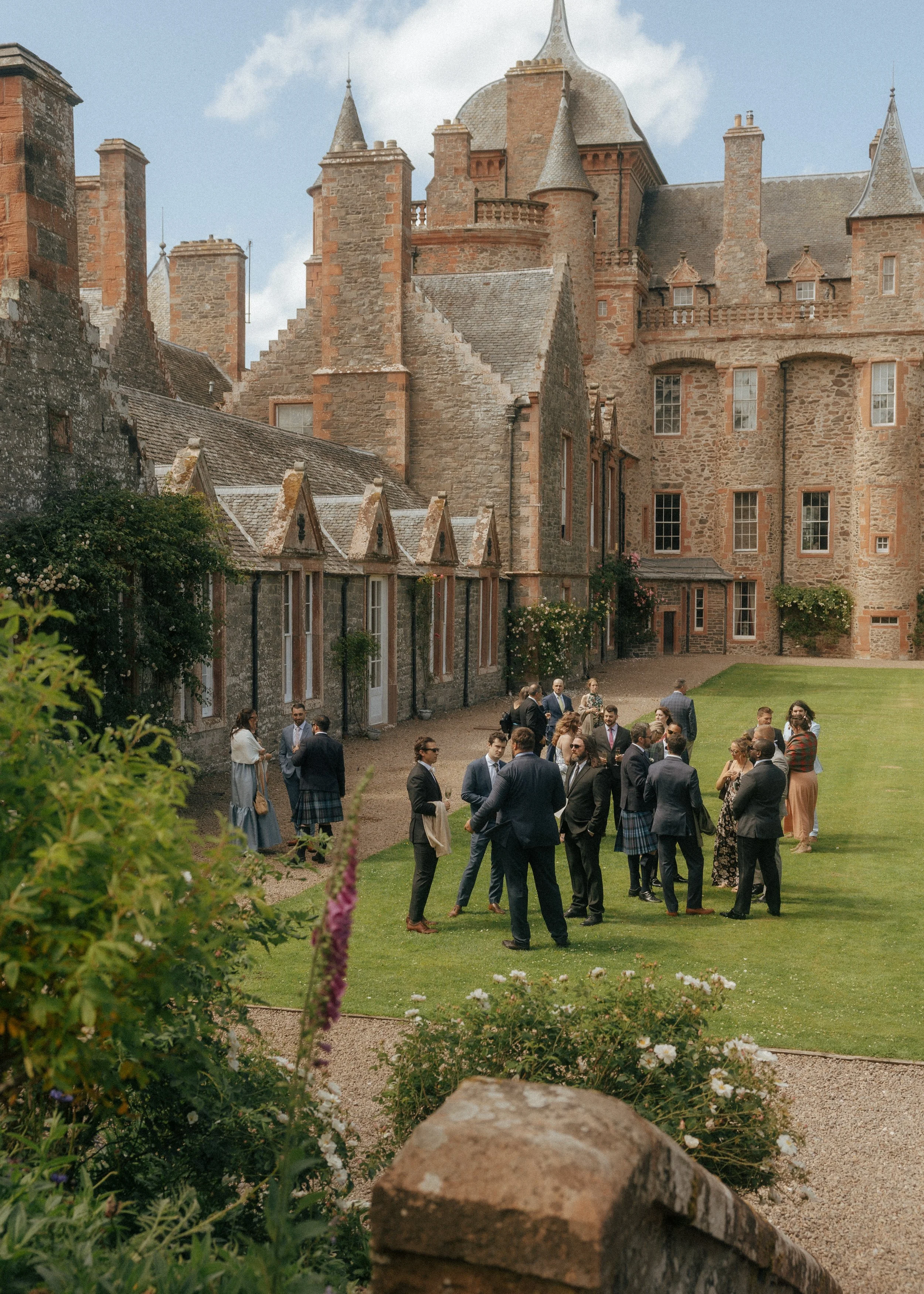 A group of people dressed in formal attire socializing in a courtyard with a historic castle in the background, surrounded by lush greenery and blooming flowers.