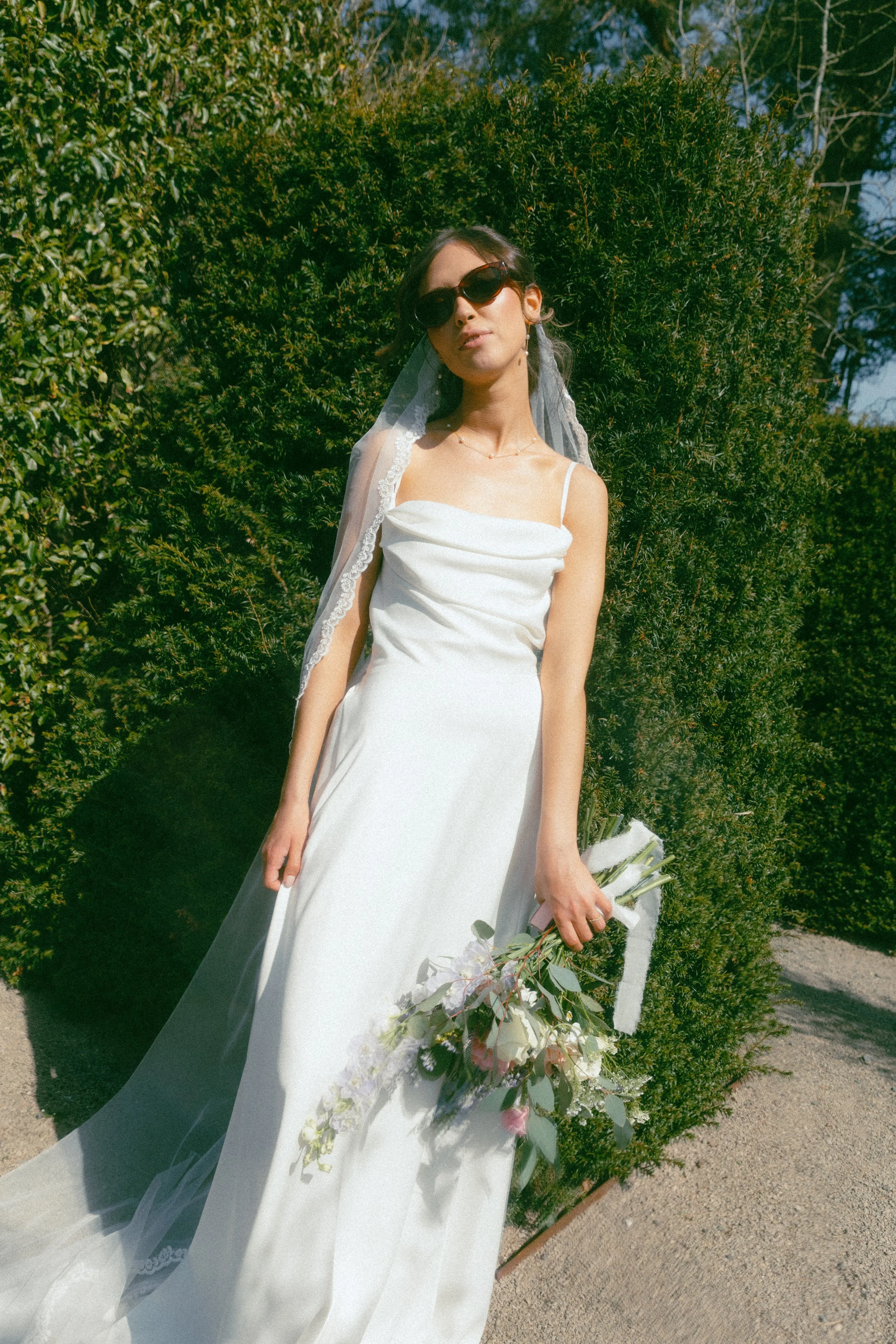A woman in a white wedding dress holding a bouquet of flowers, standing outdoors in front of green bushes, wearing sunglasses and a veil.