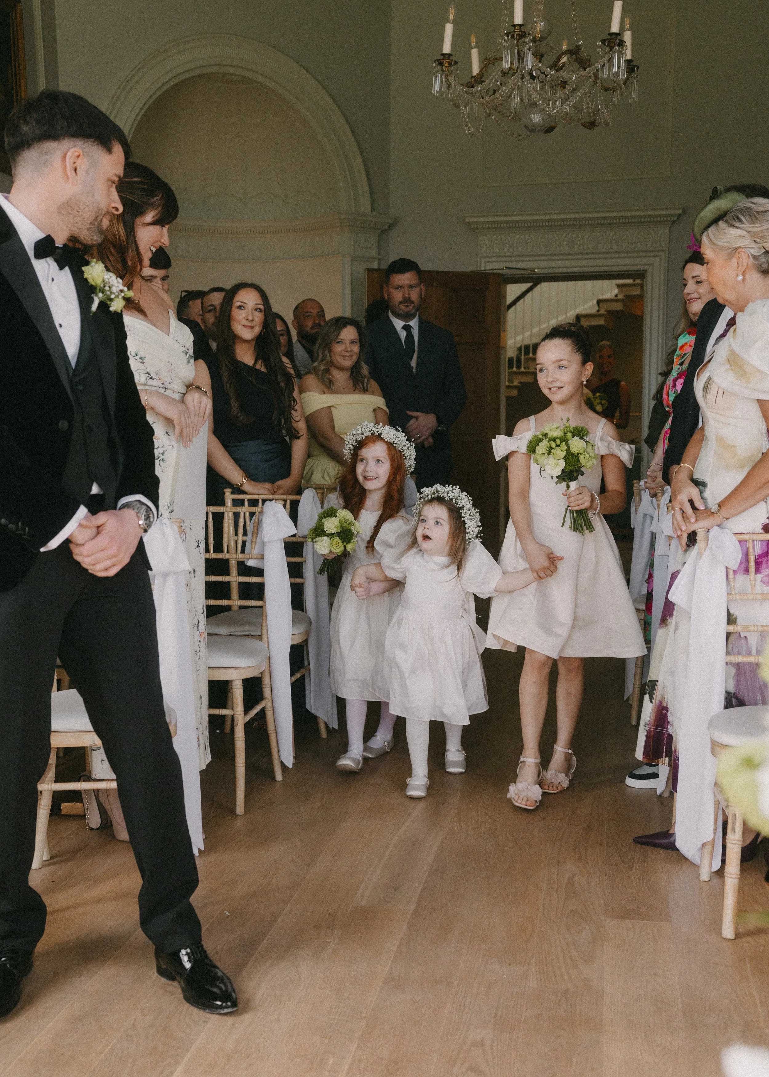 A wedding celebration with children and adults gathered in a decorated room. Two young girls in white dresses holding bouquets, and a lady in floral attire and a hat among the guests. The room has a chandelier and elegant decor.