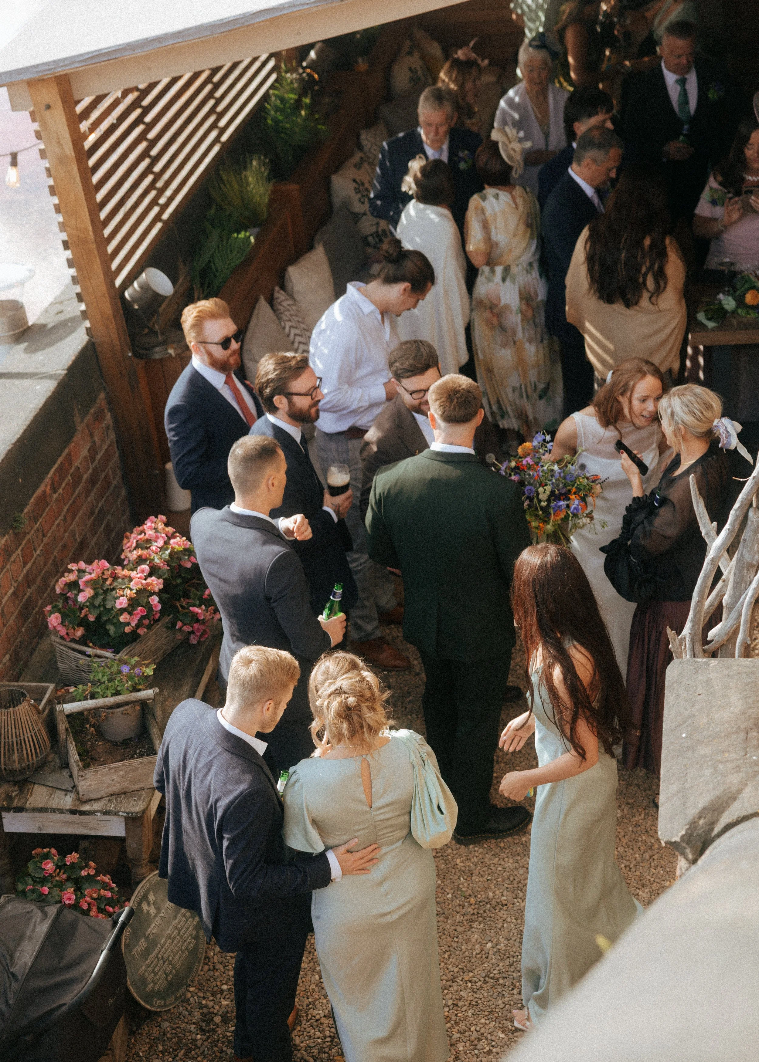 A group of people at a wedding, including women in dresses, men in suits, and a pregnant woman in a light-colored dress, gathered on a patio with flower arrangements and a stroller nearby.