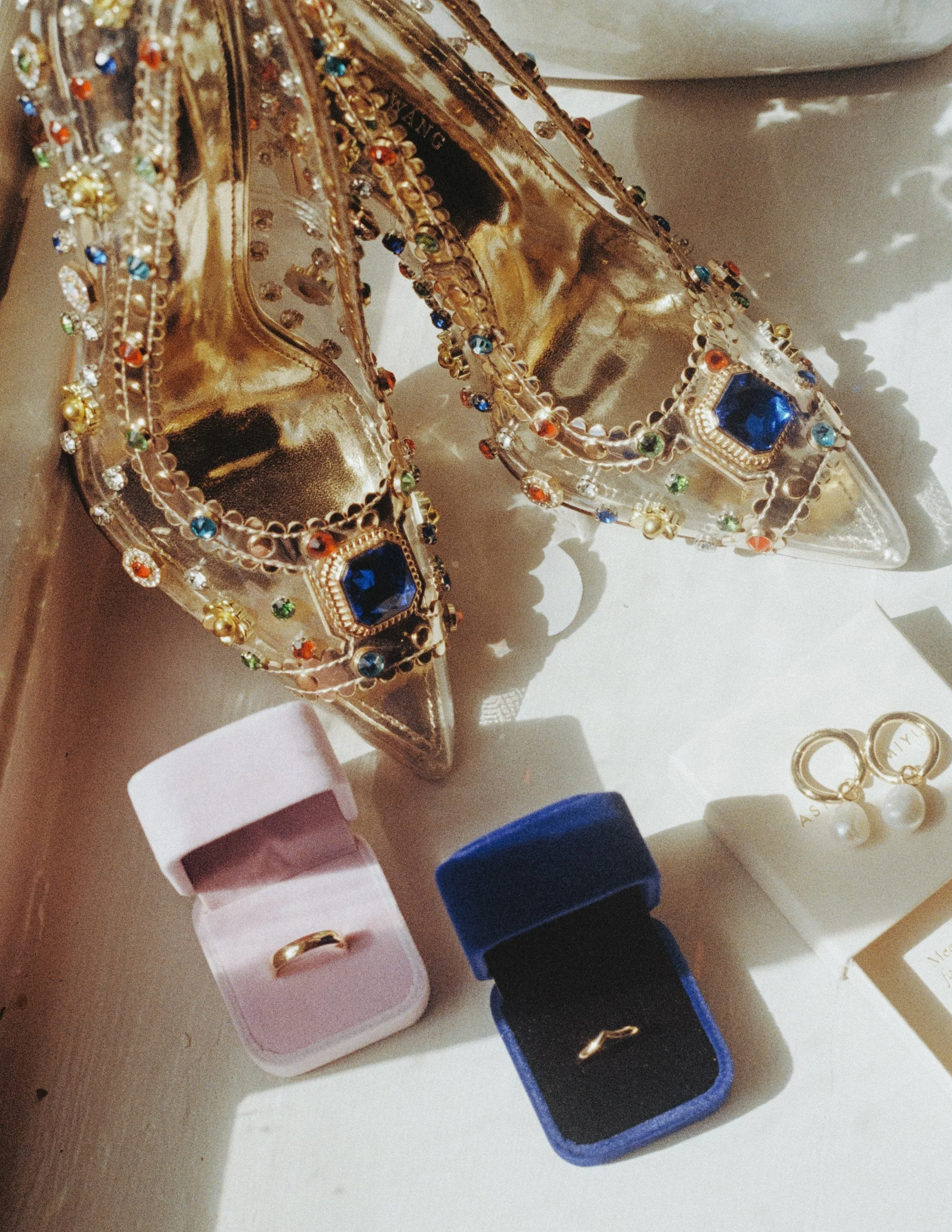 Gold high-heeled shoes decorated with colorful rhinestones and large blue gemstones, accompanied by jewelry including pearl earrings and rings in boxes.