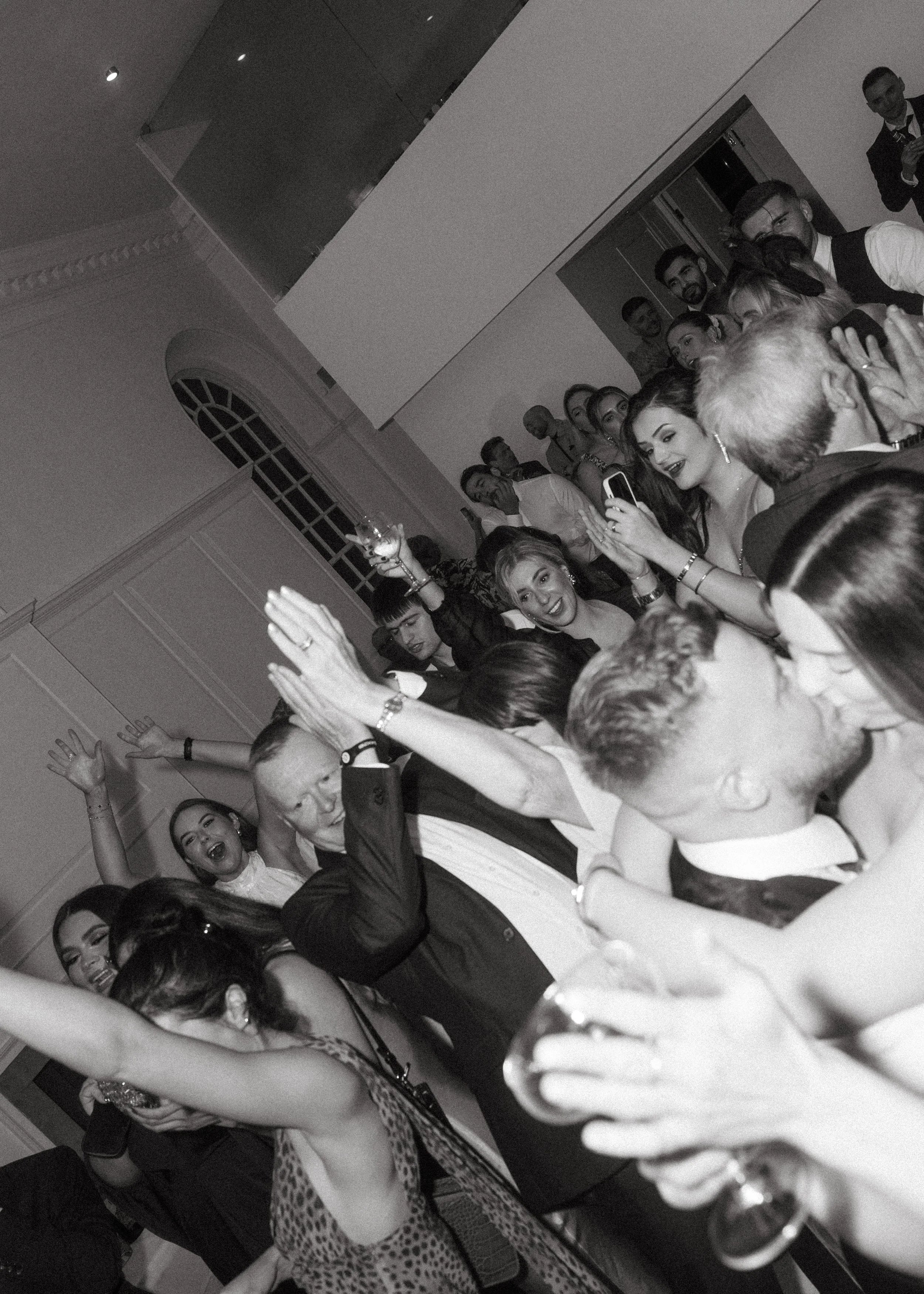 Group of people dancing and celebrating at a party, some are holding glasses and taking selfies, in an elegant indoor setting.