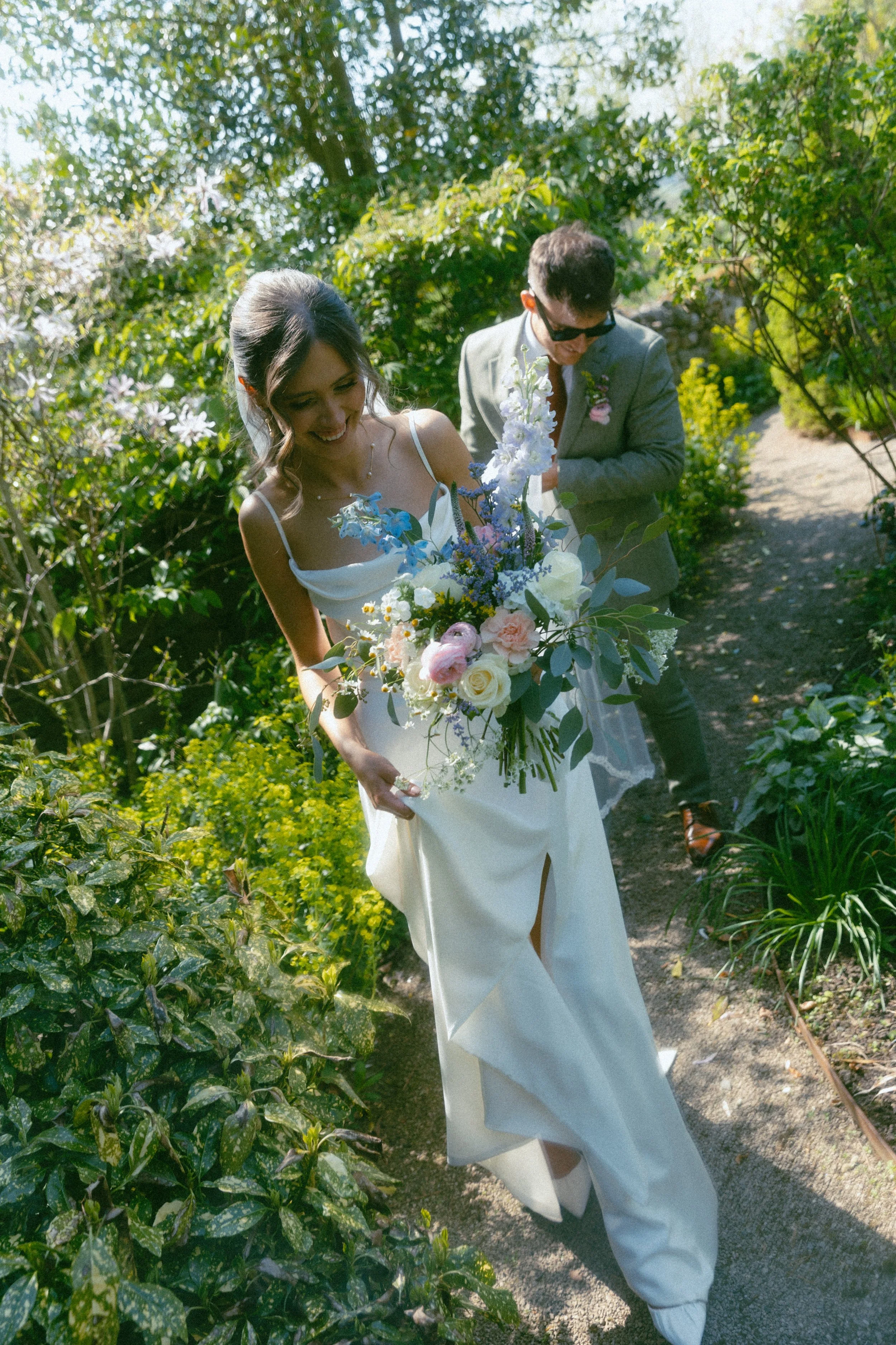 A bride in a white wedding dress holding a bouquet of flowers, walking on a garden path with a groom in a light gray suit and sunglasses behind her.
