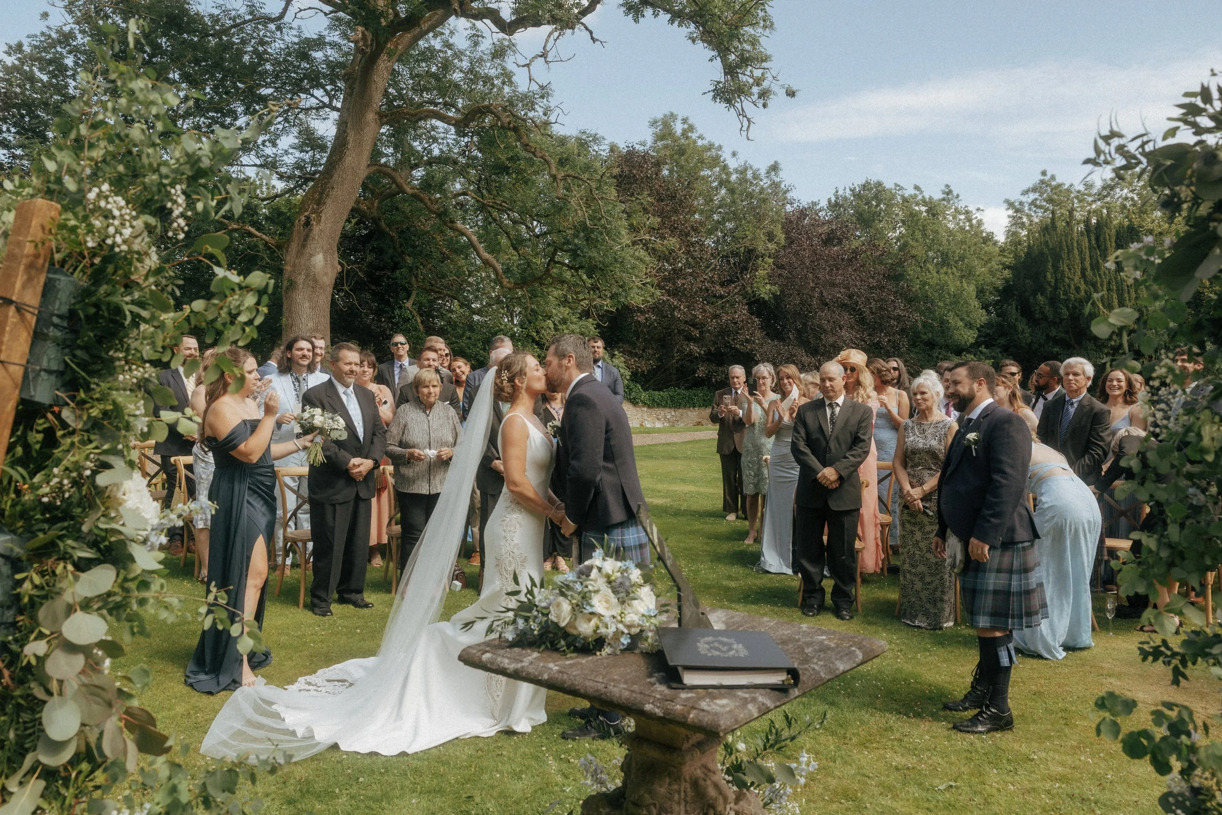 A wedding ceremony outdoors with guests, a bride and groom kissing, surrounded by trees and greenery.