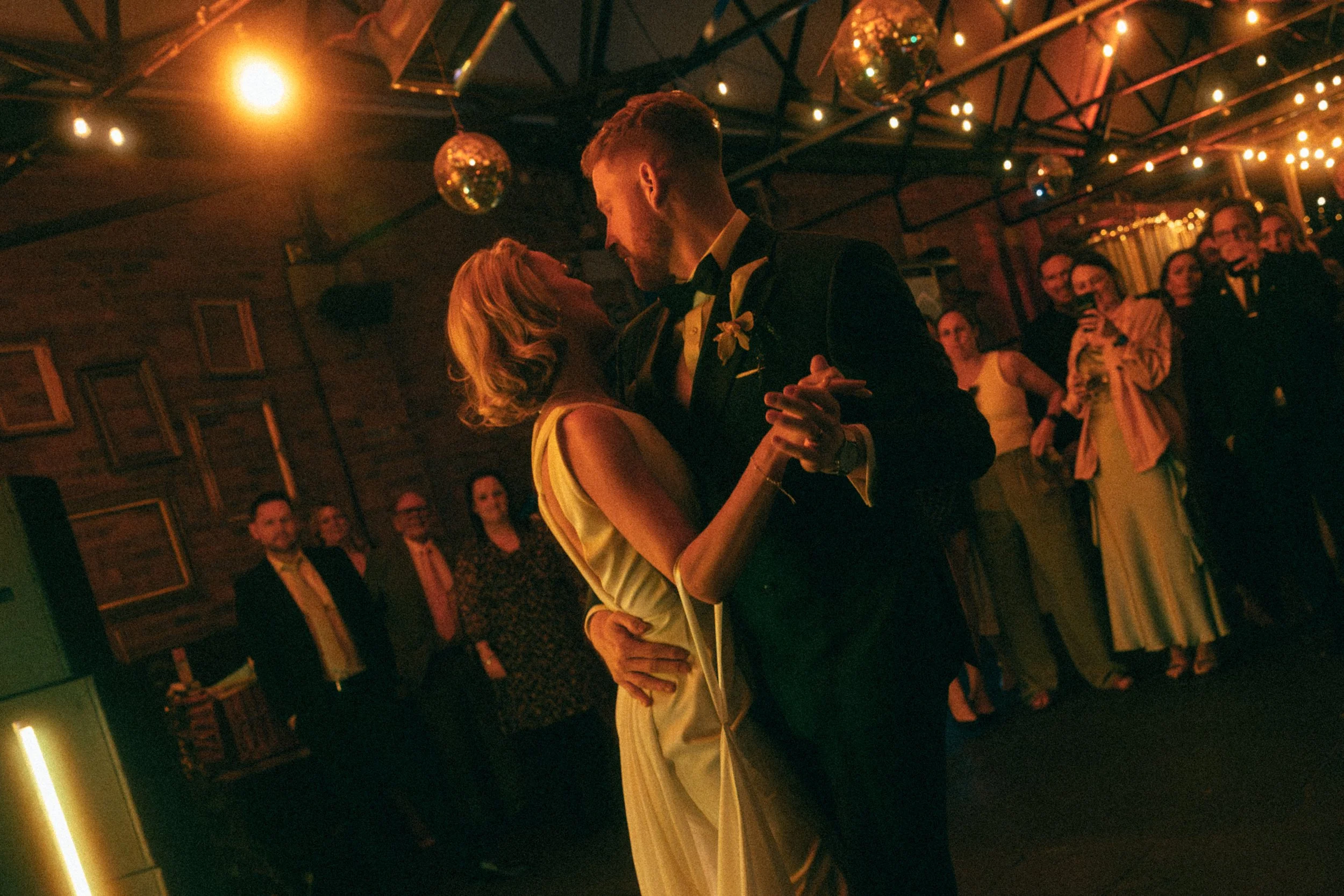A couple dancing closely at a party with string lights and disco balls hanging from ceiling, surrounded by onlookers.