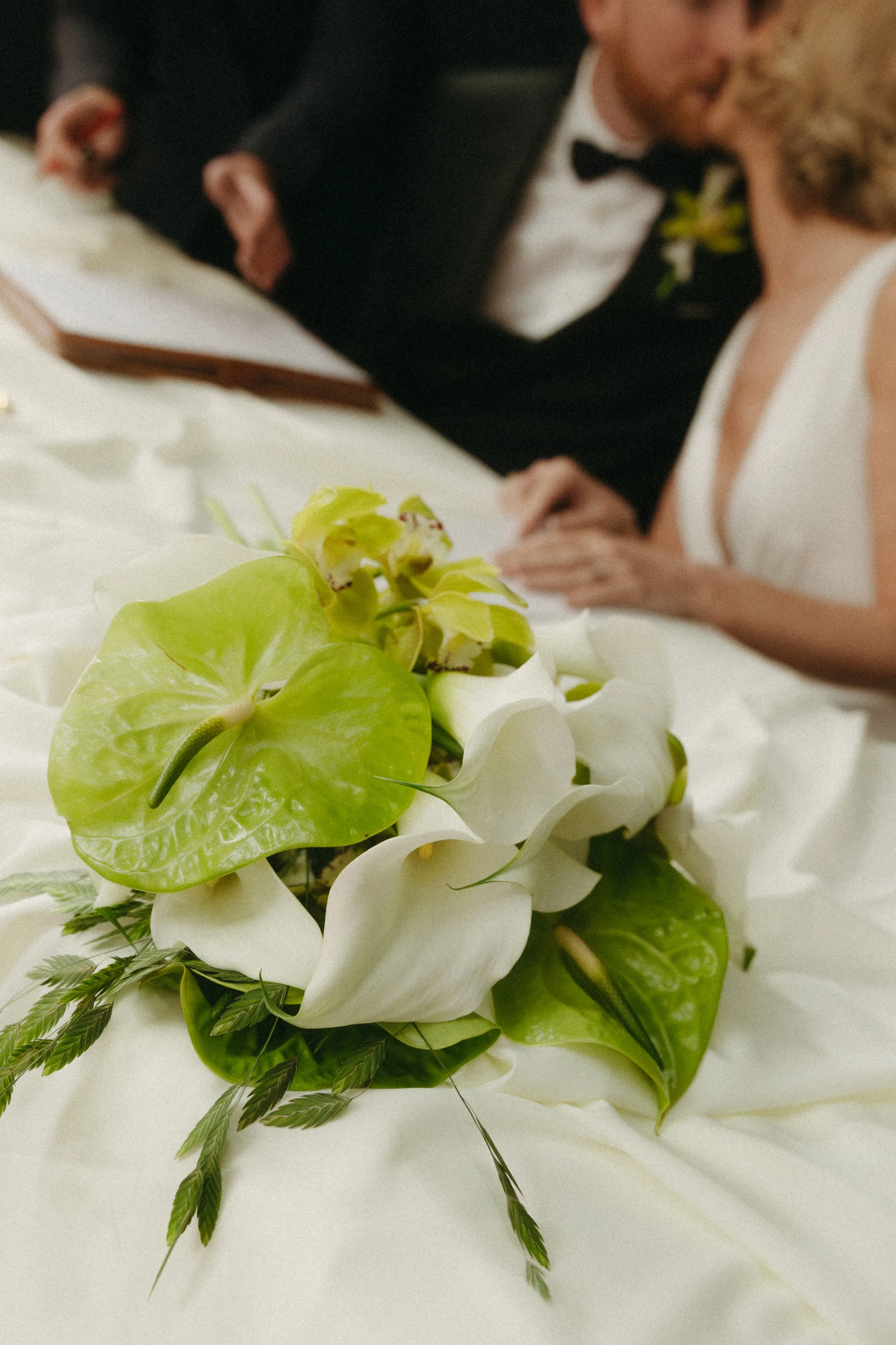 Close-up of a wedding bouquet with white flowers, green leaves, and yellow-green accents, lying on a white surface, with a bride and groom in the background kissing.
