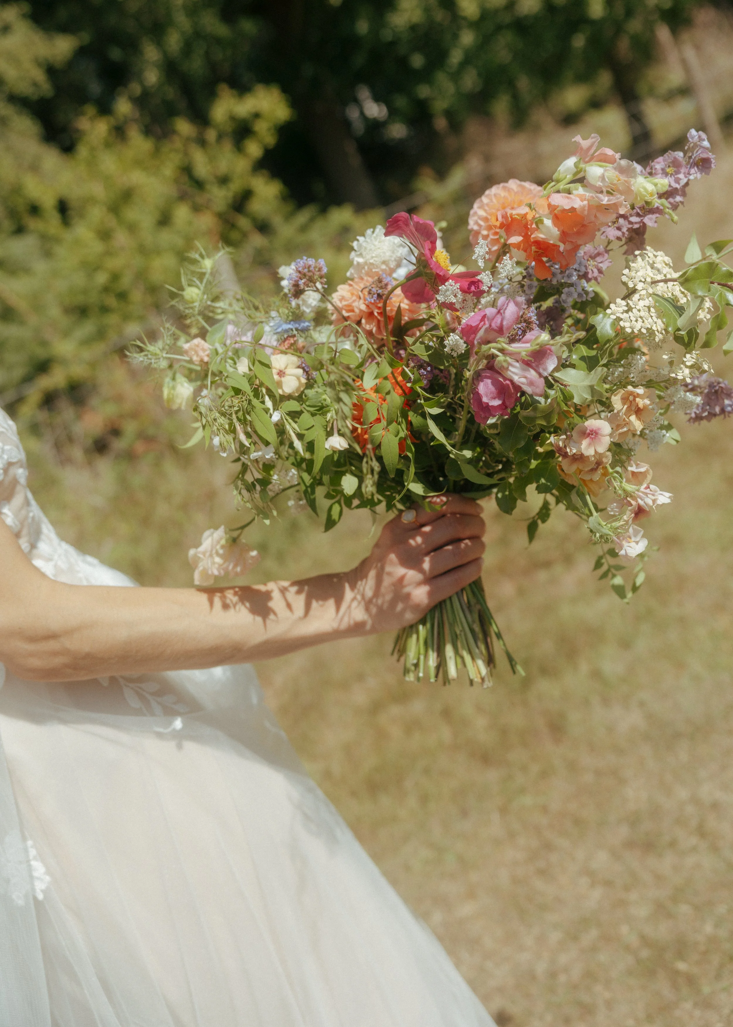 A woman dressed in a white wedding dress holds a large bouquet of mixed flowers outdoors in a sunny setting with green trees in the background.