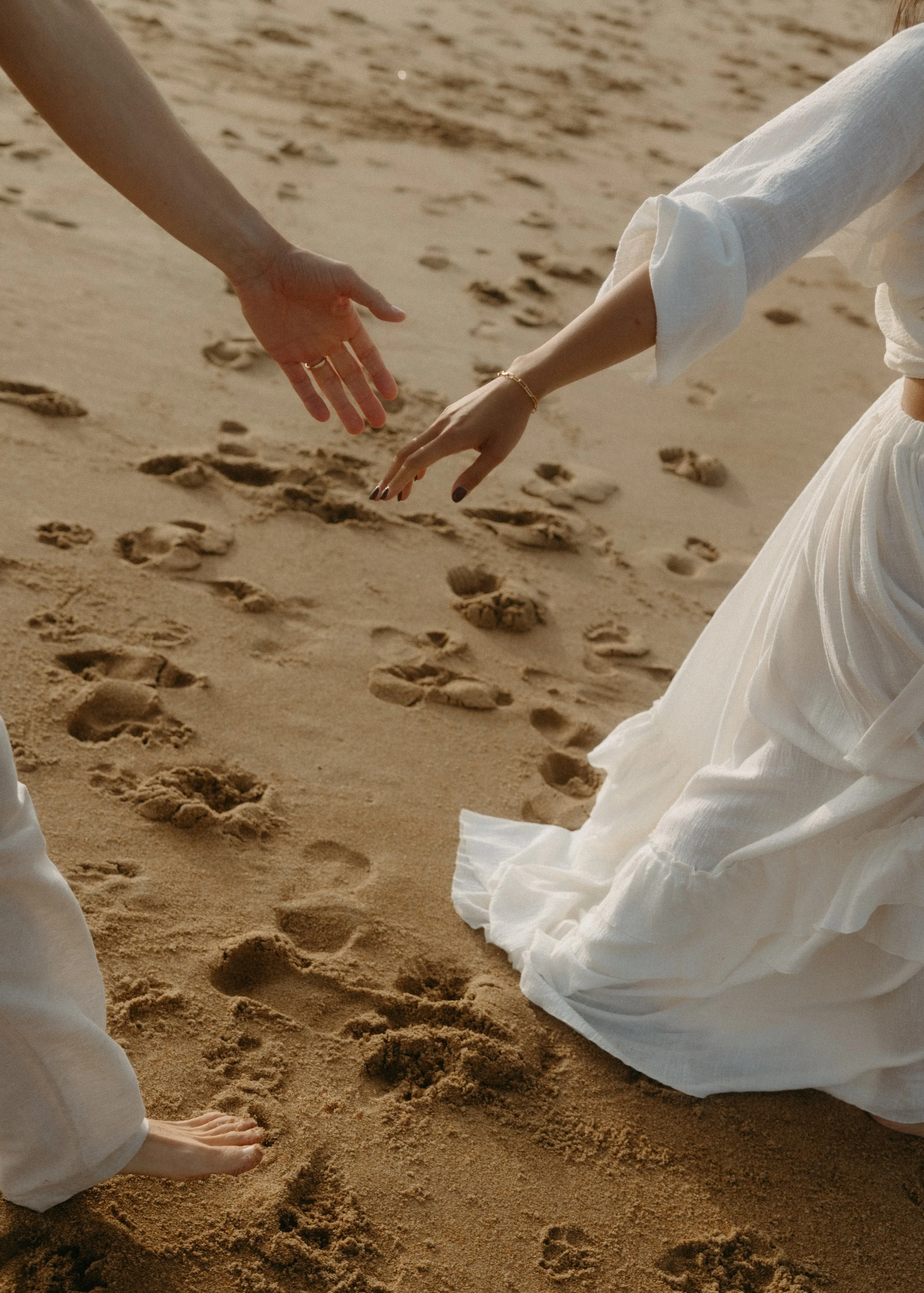 Two people reaching towards each other on a sandy beach with footprints, one in a white dress and the other in light-colored clothing.