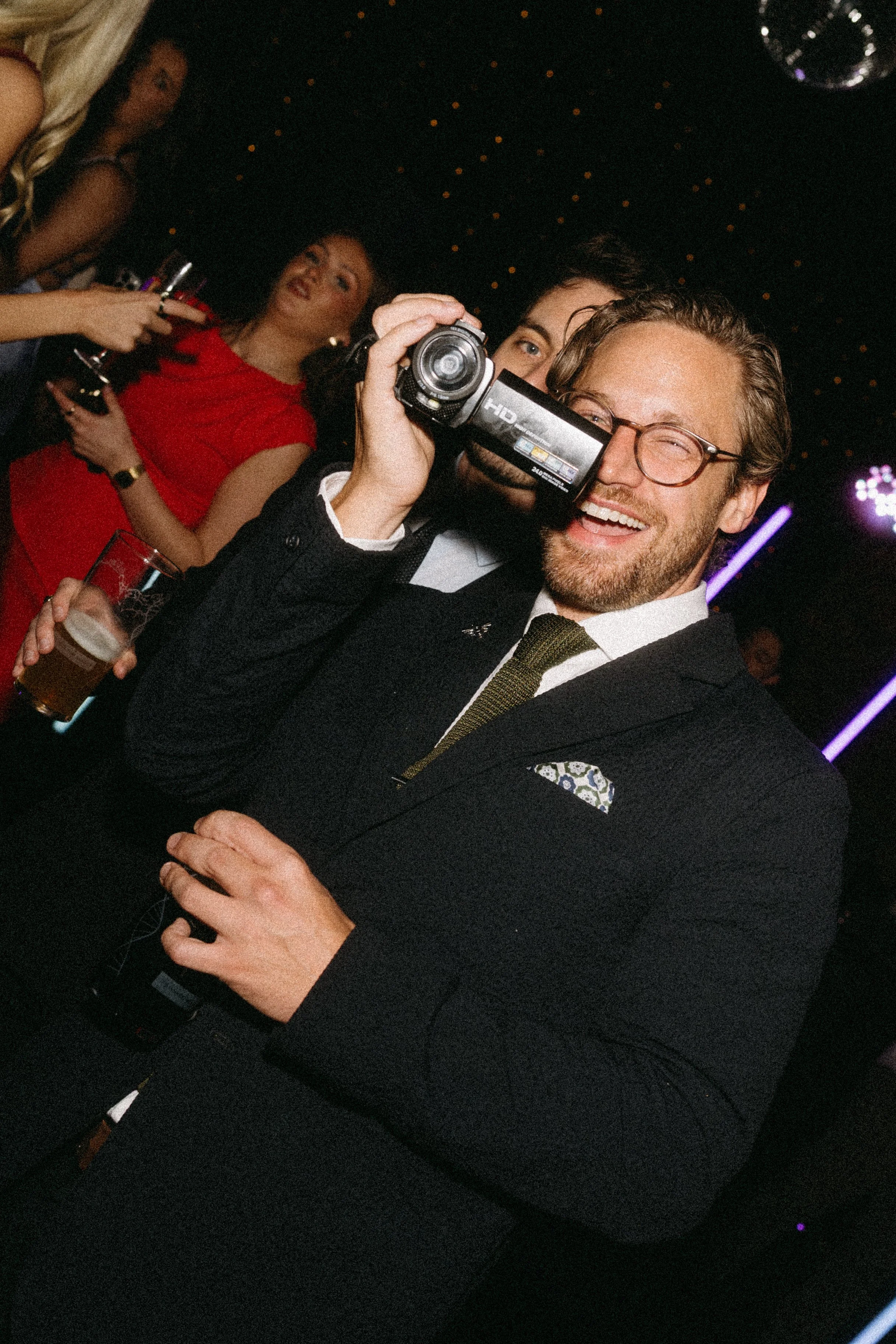 People dancing at a party with some holding drinks, a woman in a red dress, a man with glasses smiling, and festive lighting in the background.