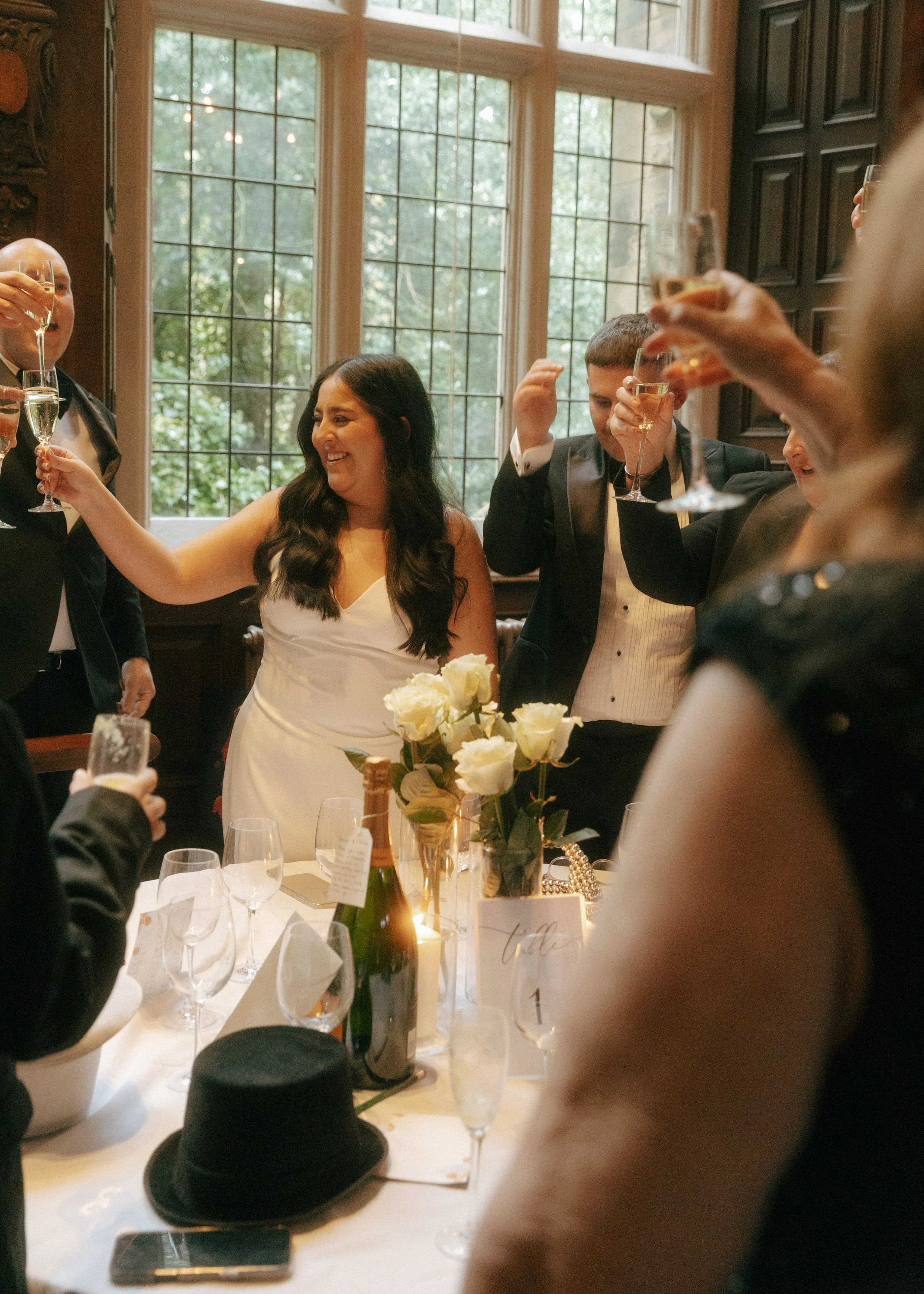 A group of people dressed in formal attire raising glasses for a toast at a celebration, with a woman in a white dress in the center holding a bouquet of flowers and smiling.