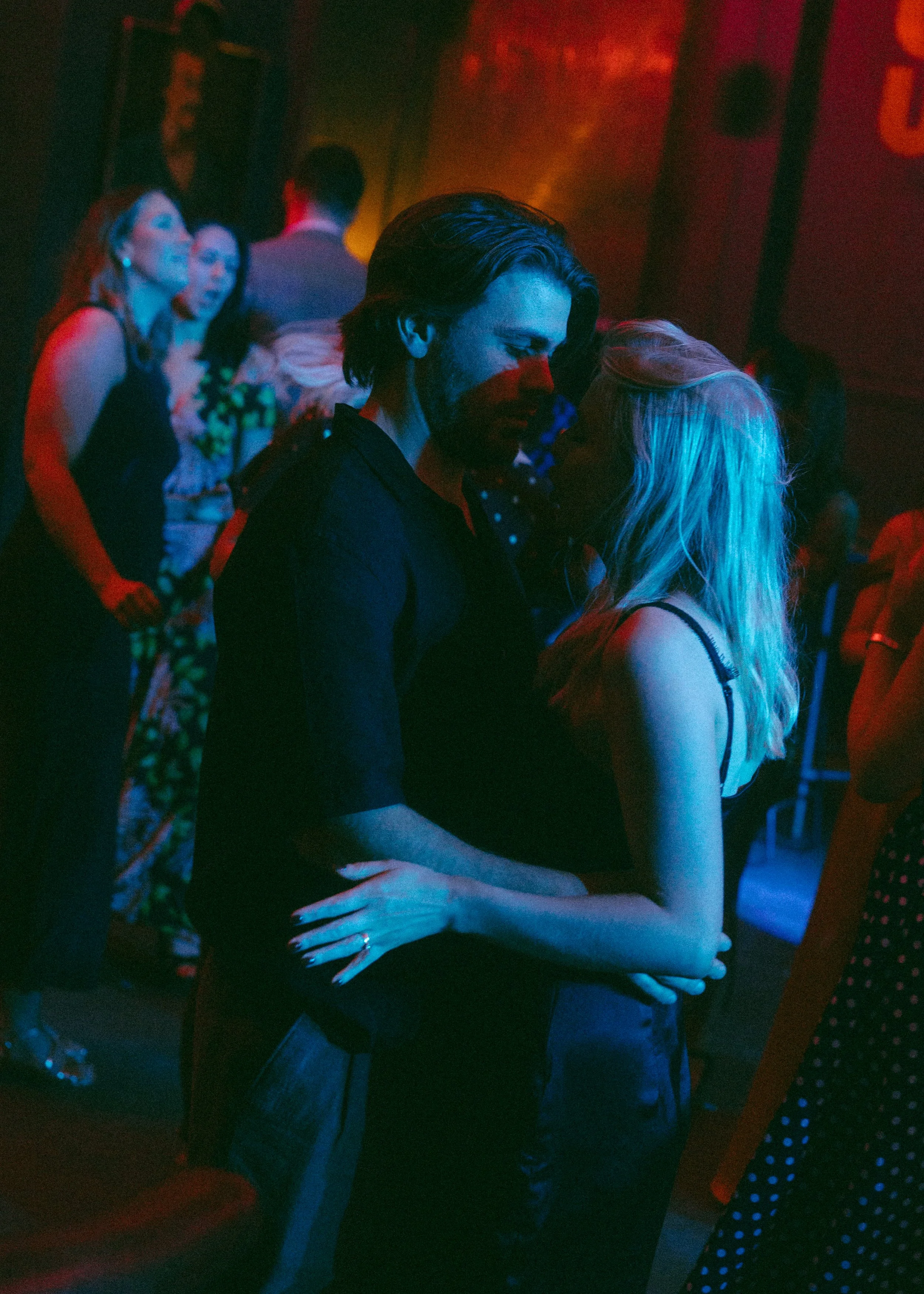 A man and a woman are dancing closely in a dimly lit room with colorful lighting, surrounded by other people who are also dancing and socializing.