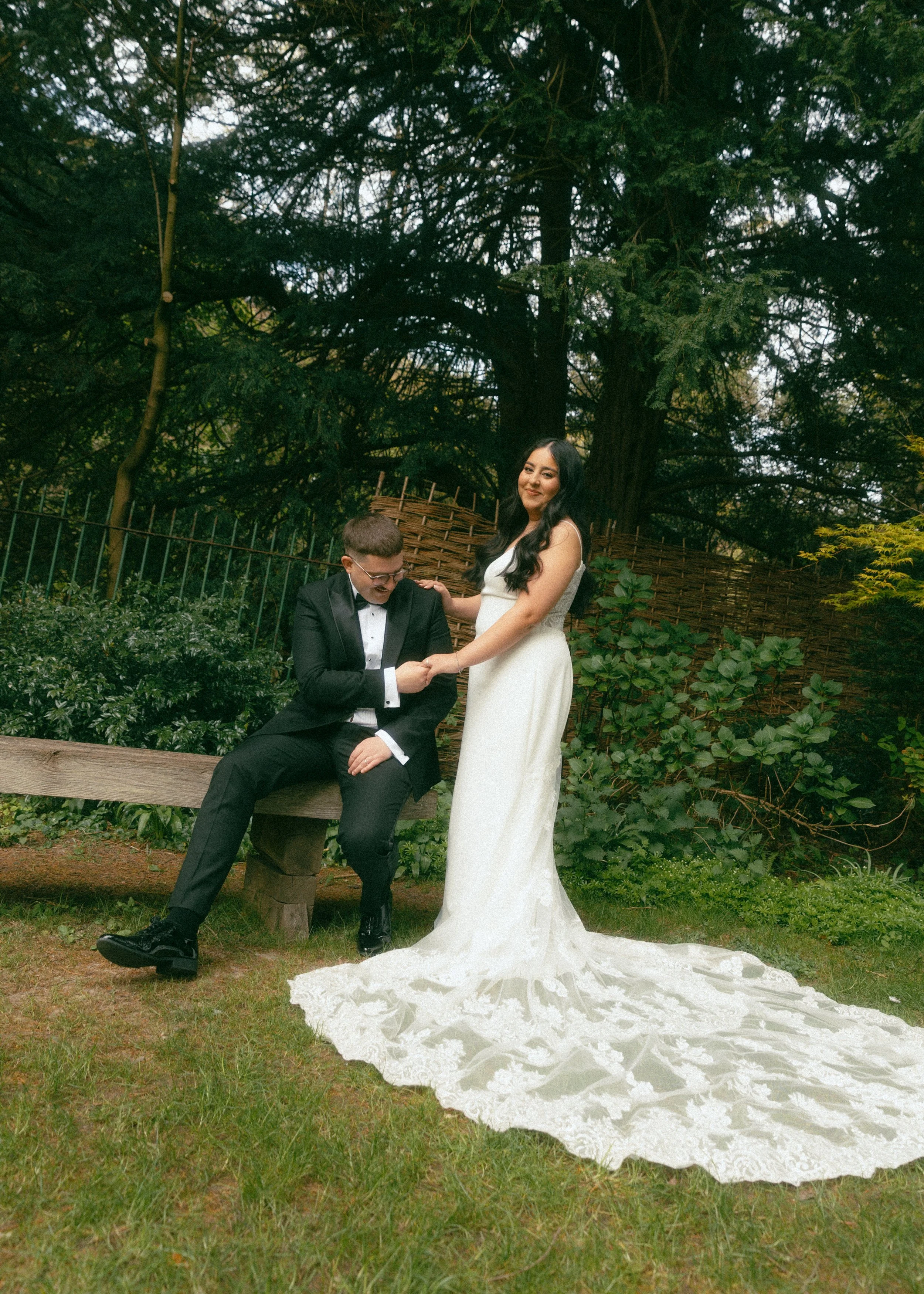 A bride and groom are outdoors in a garden-like setting. The bride is in a white wedding dress, standing, smiling, and touching the groom's hand. The groom, dressed in a black tuxedo and glasses, is sitting on a bench, looking down, with her hand on 