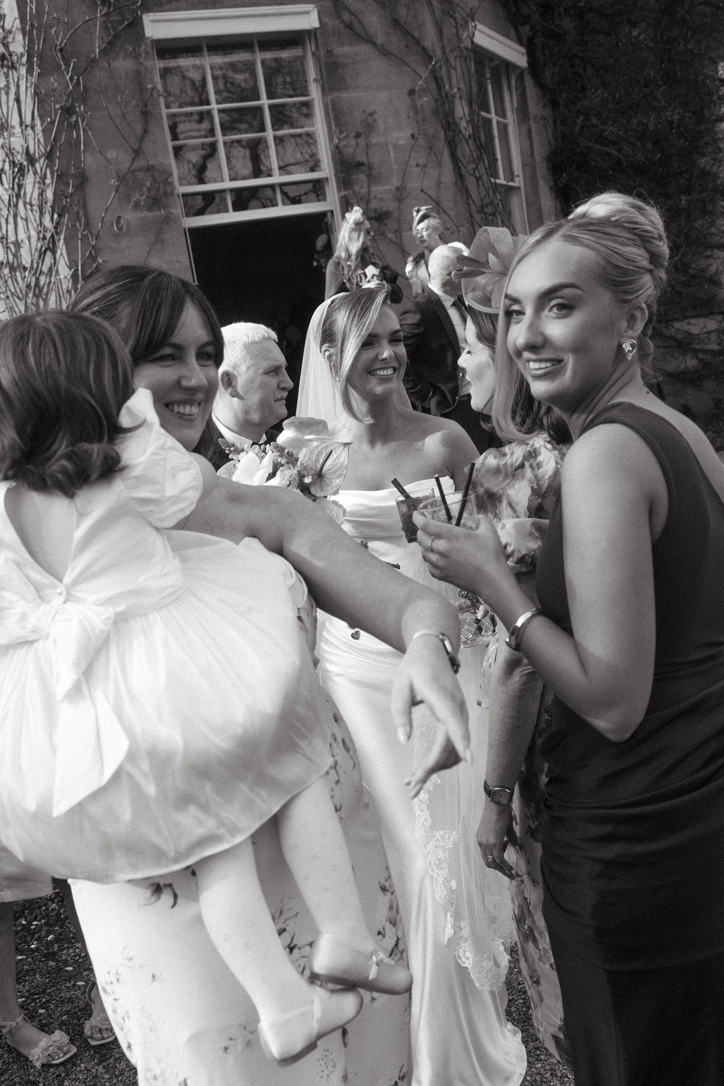Black and white photo of a group of women at a wedding, talking and smiling. The bride is in the center, wearing a wedding gown and veil, holding a bouquet. Other women are dressed in various dresses, some with floral patterns, and holding drinks. Th