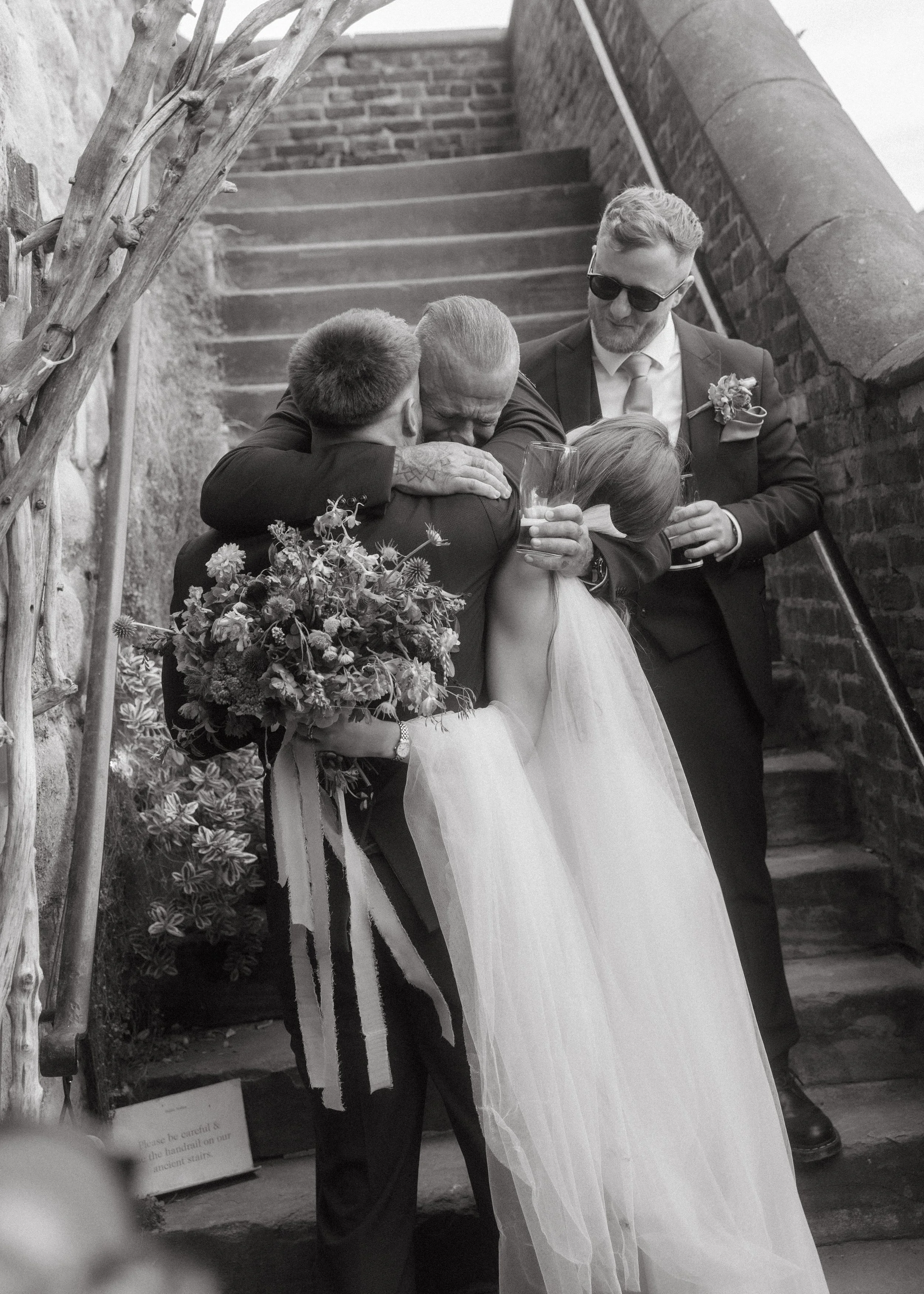 A black and white photo of a wedding scene showing a bride in a white dress hugging an older man, possibly her father, with a man in a suit and sunglasses standing nearby. They are outside near brick stairs and a brick wall.