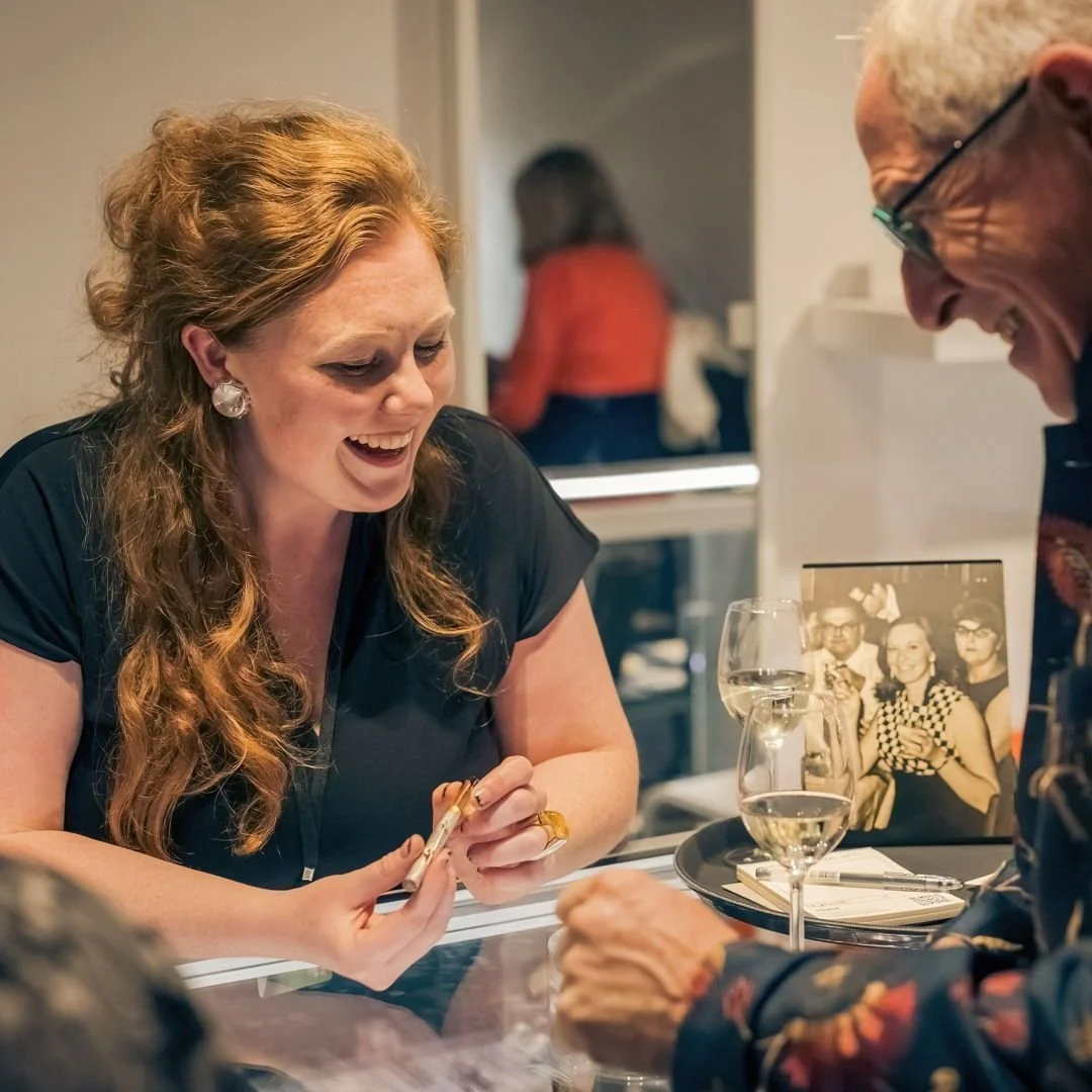 A woman with long, curly red hair and beard, smiling and looking at a small item she is holding, while a man with glasses and white hair also smiles. They are sitting at a table with wine glasses and a black tray, and there is a vintage photo on the table showing two women and a man.