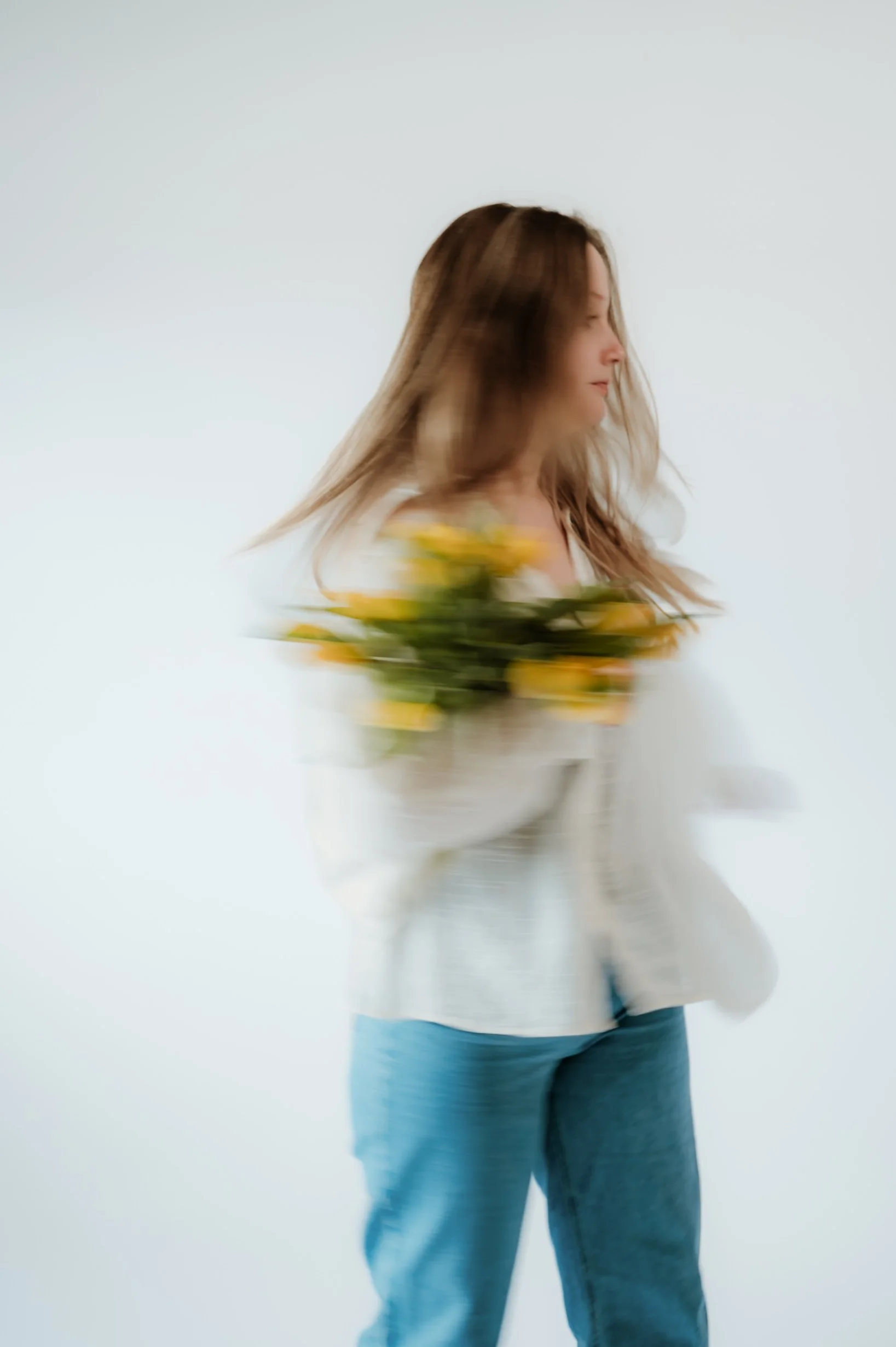 A woman with brown hair is holding a bouquet of yellow flowers and walking, wearing a white top and blue jeans, against a plain white background.