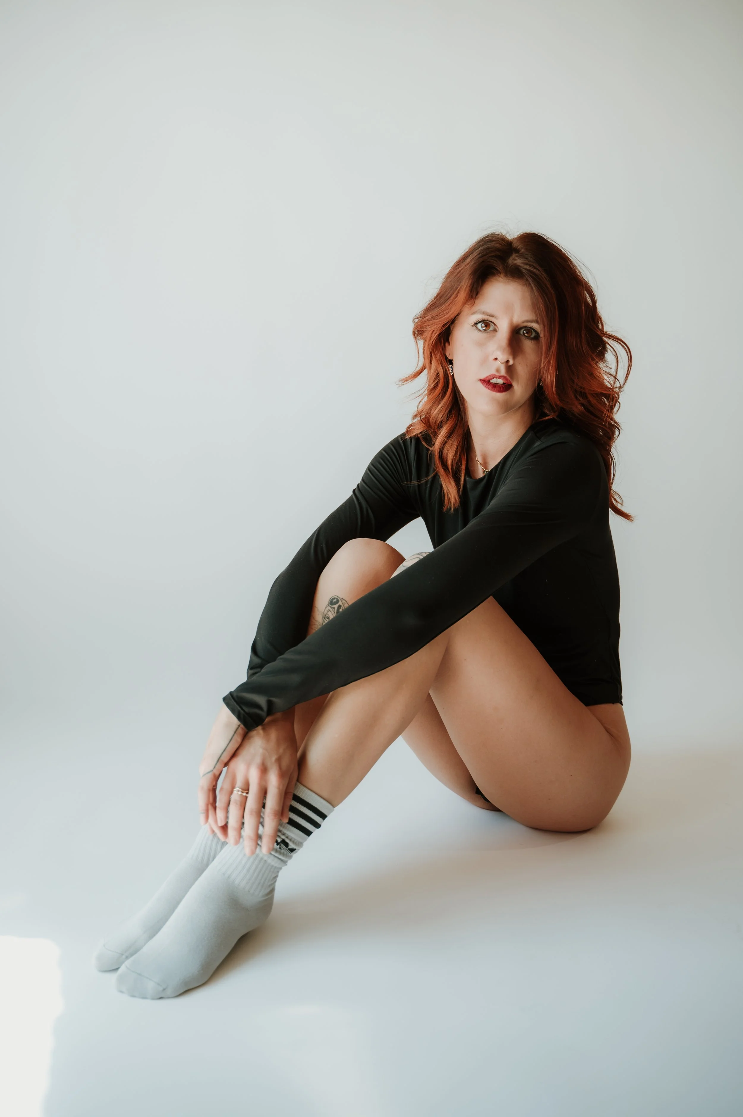 A young woman with red hair and fair skin, wearing a black long-sleeve shirt, striped socks, and white shoes, sitting against a plain light gray background with her knees pulled to her chest and looking at the camera.