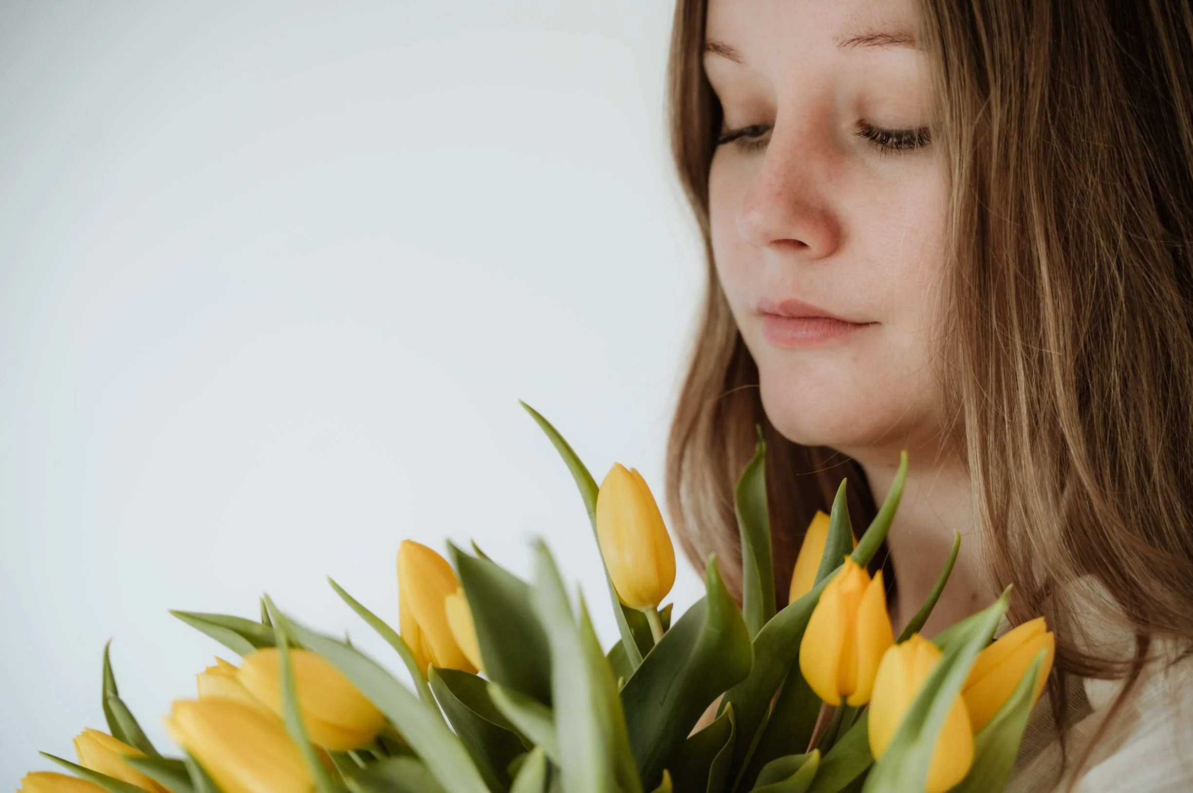 A woman with light skin and long, light brown hair holding a bouquet of yellow tulips. She has her eyes closed and a peaceful expression on her face.