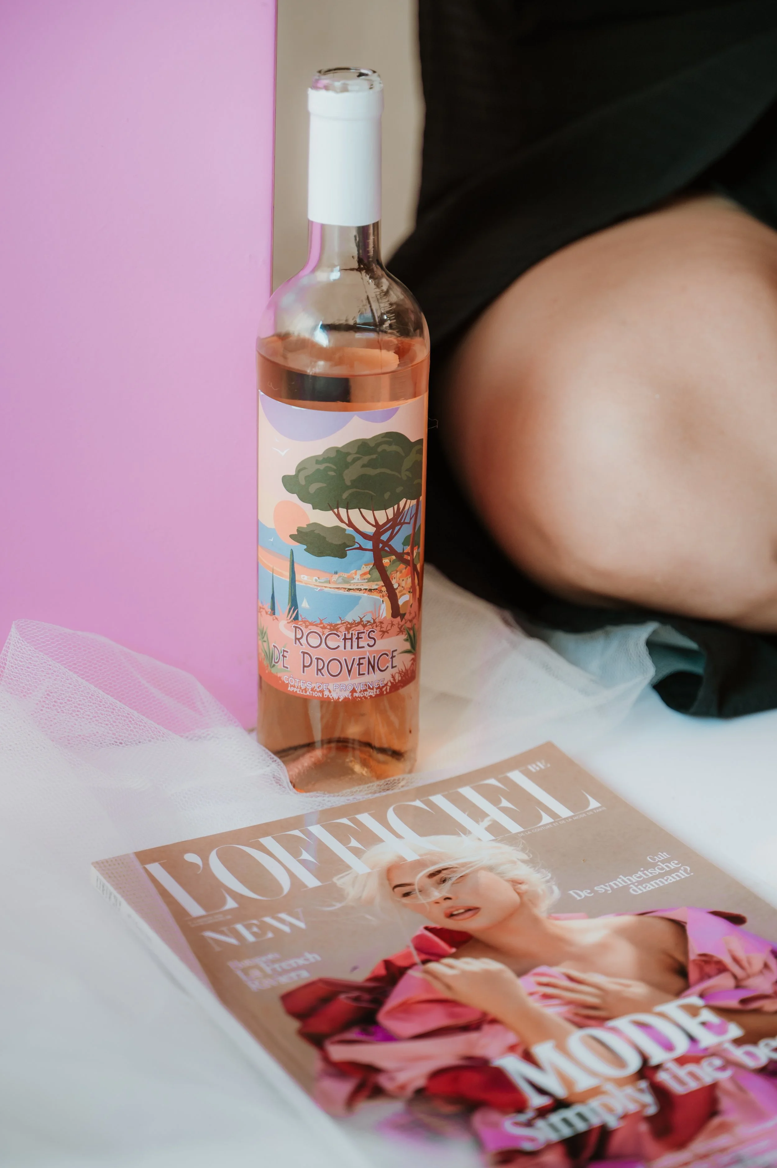 A bottle of rosé wine named 'Roches de Provence' with scenic illustration of trees, mountains, and a sunset, positioned next to a pink box and a La Vie magazine on a white surface, with a person's arm and black shirt partially visible in the backgrou