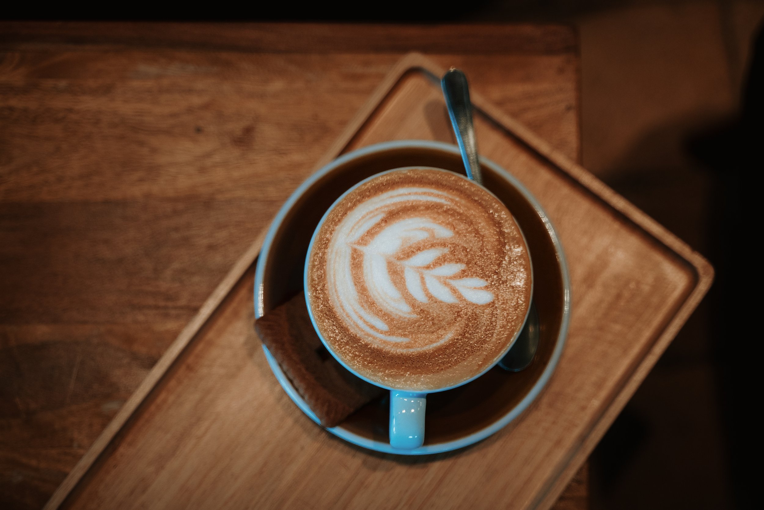 A cup of coffee with latte art resembling a leaf, placed on a wooden surface with a small spoon inside the cup.