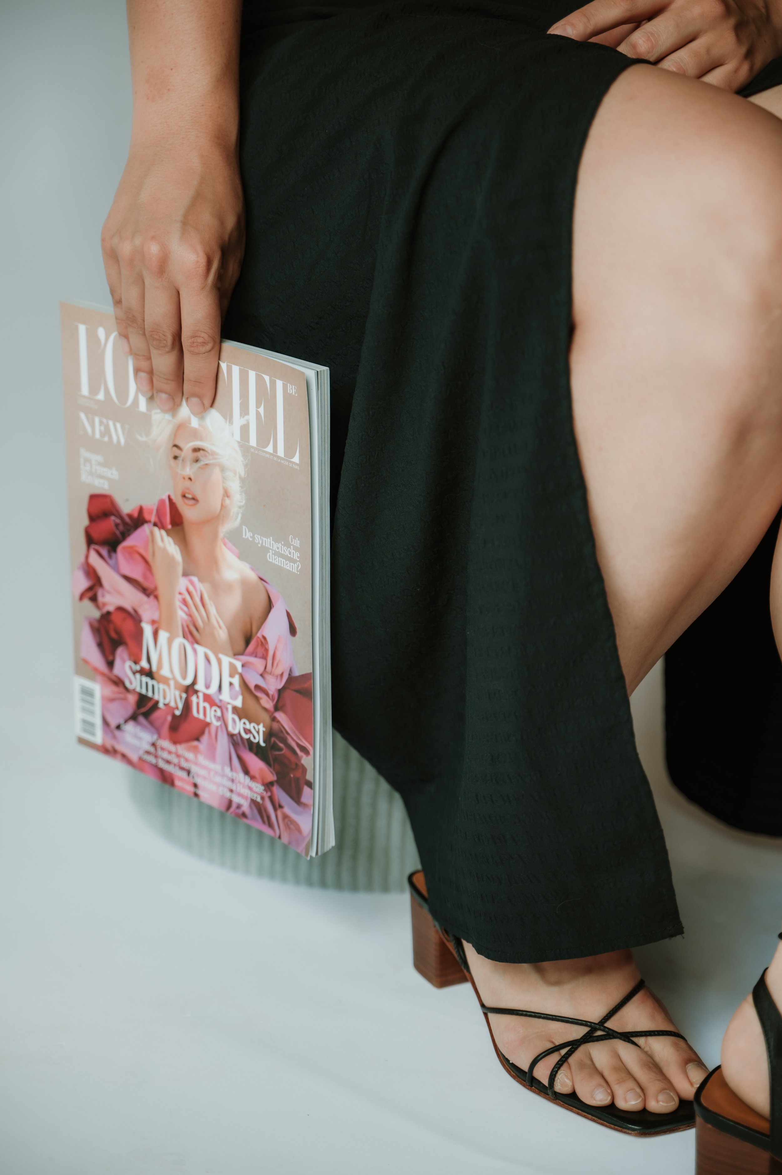 Close-up of a woman's hand holding a fashion magazine, her arm resting on a black dress, with her feet in black strappy sandals and a wooden block heel visible.