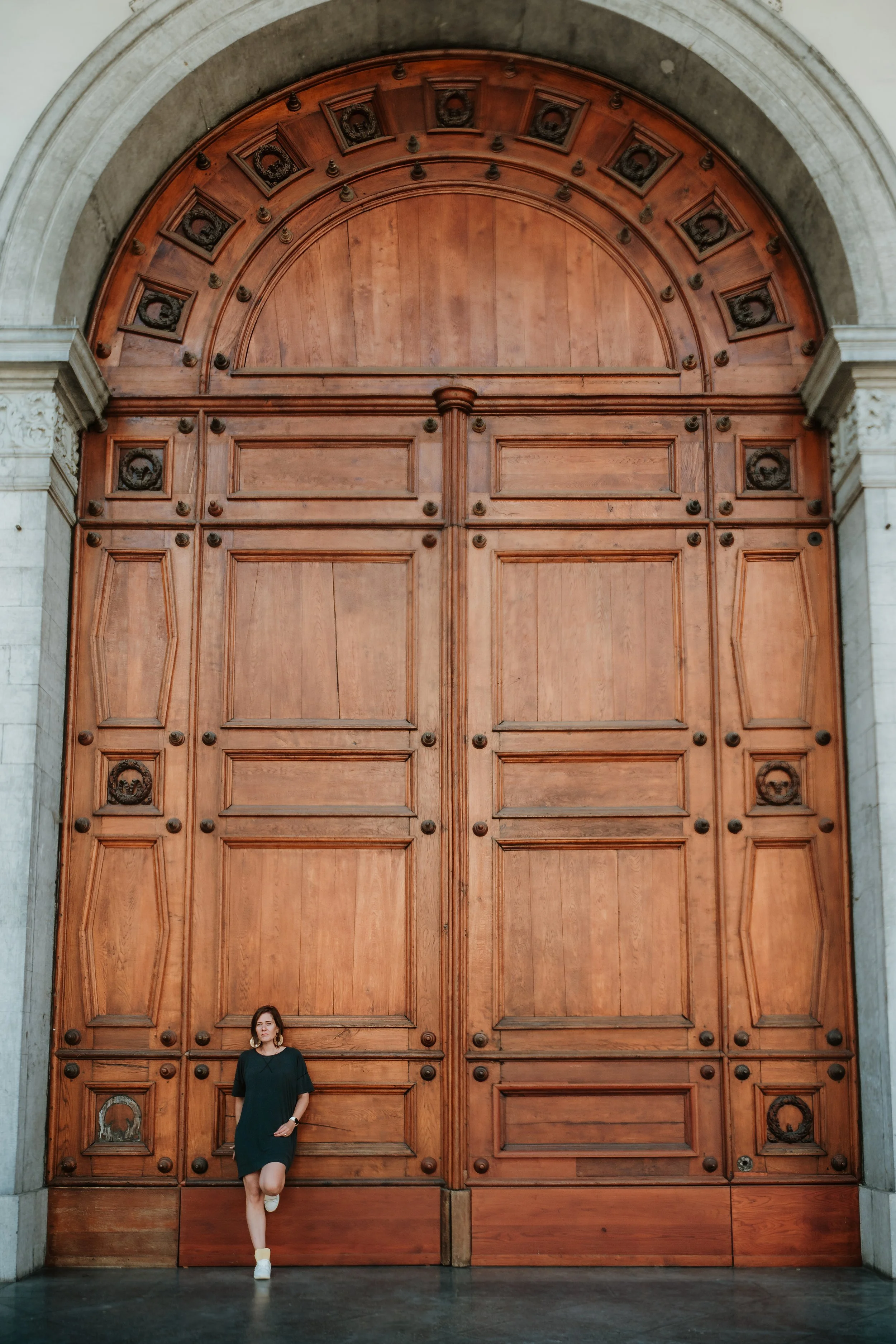 A woman in a dark green dress and white shoes standing in front of an ornate large wooden door with decorative panels and metal accents, part of an architectural structure with stone columns.