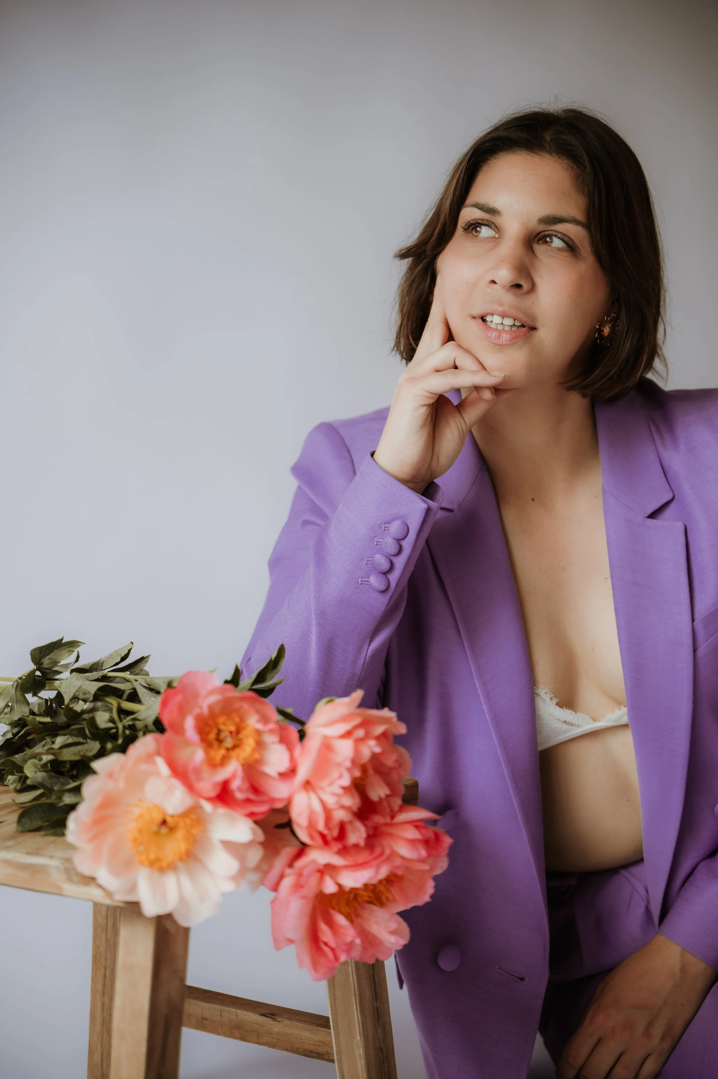A woman in a purple blazer sitting next to a wooden stand with pink and orange flowers on it, looking thoughtfully to the right.
