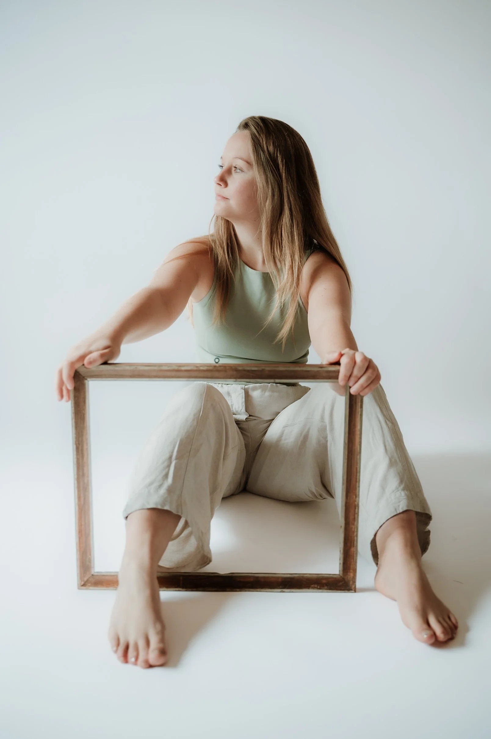 A woman sitting on the floor, holding a wooden frame, with her legs extended and her head turned to the side, against a plain light-colored background.