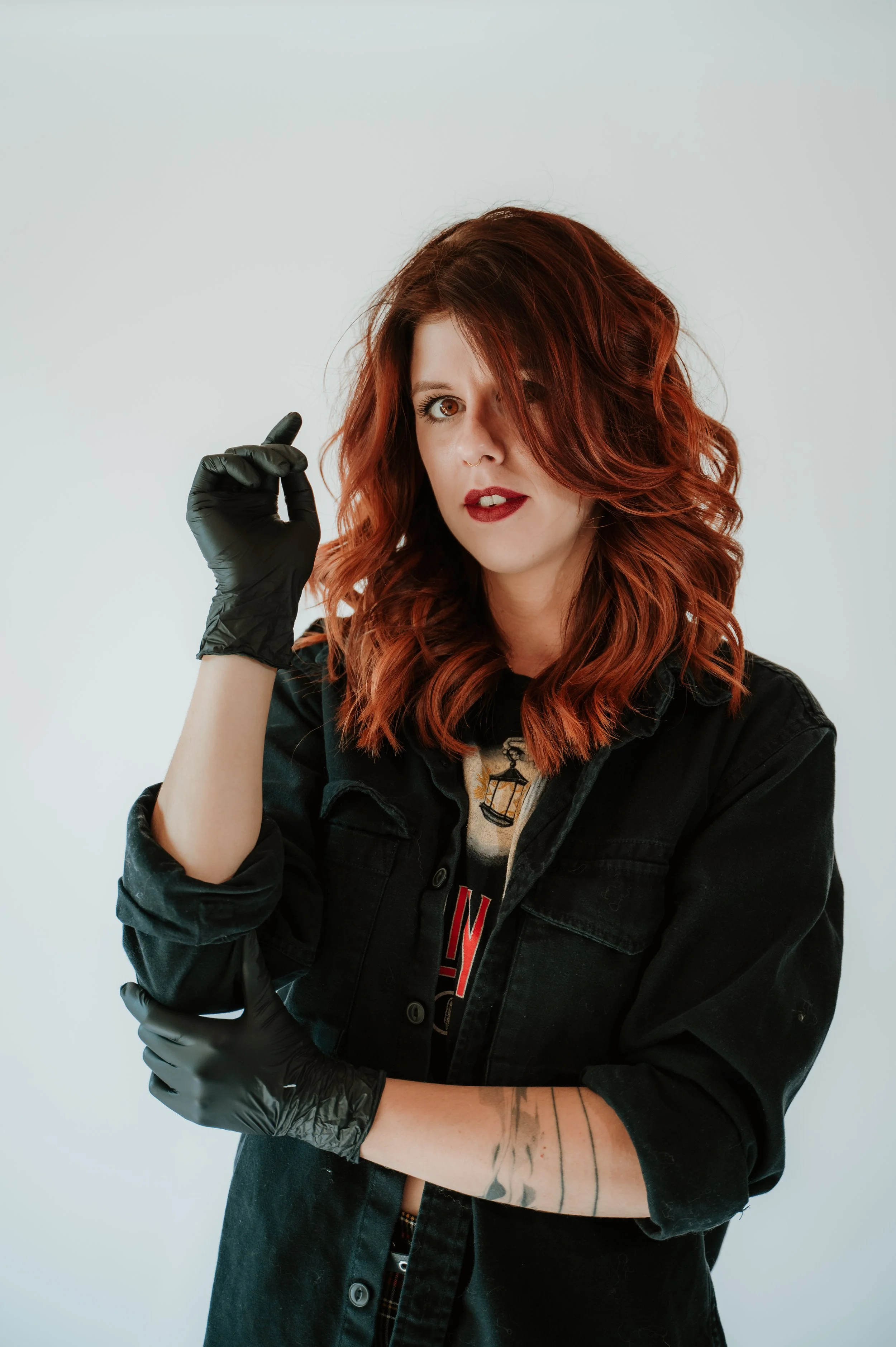 A woman with wavy red hair and brown eyes wearing a black denim jacket, black gloves, and a graphic t-shirt, standing against a plain white background.