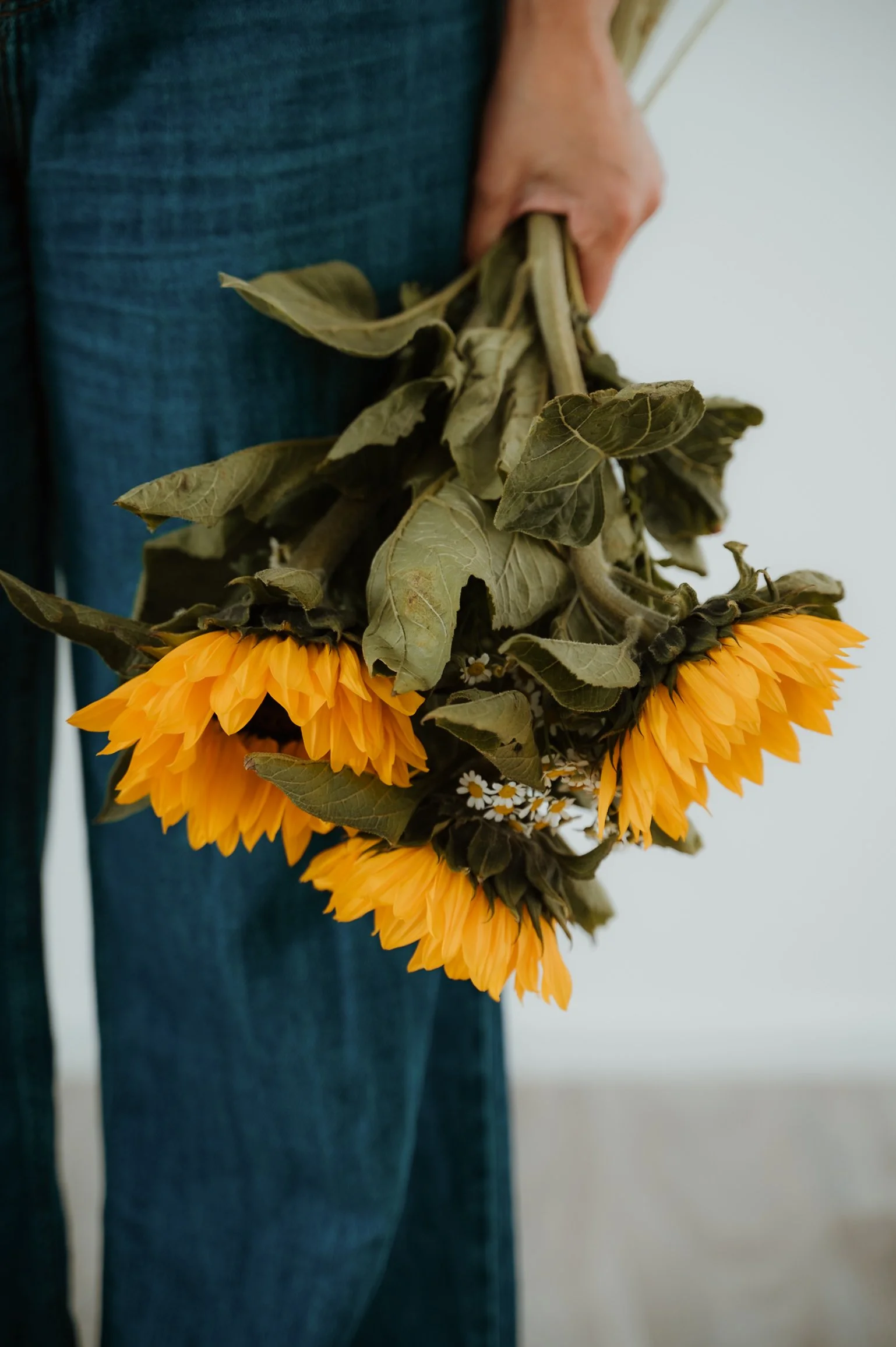 A person in blue jeans holding a bouquet of sunflowers and dried leaves against a plain light background.