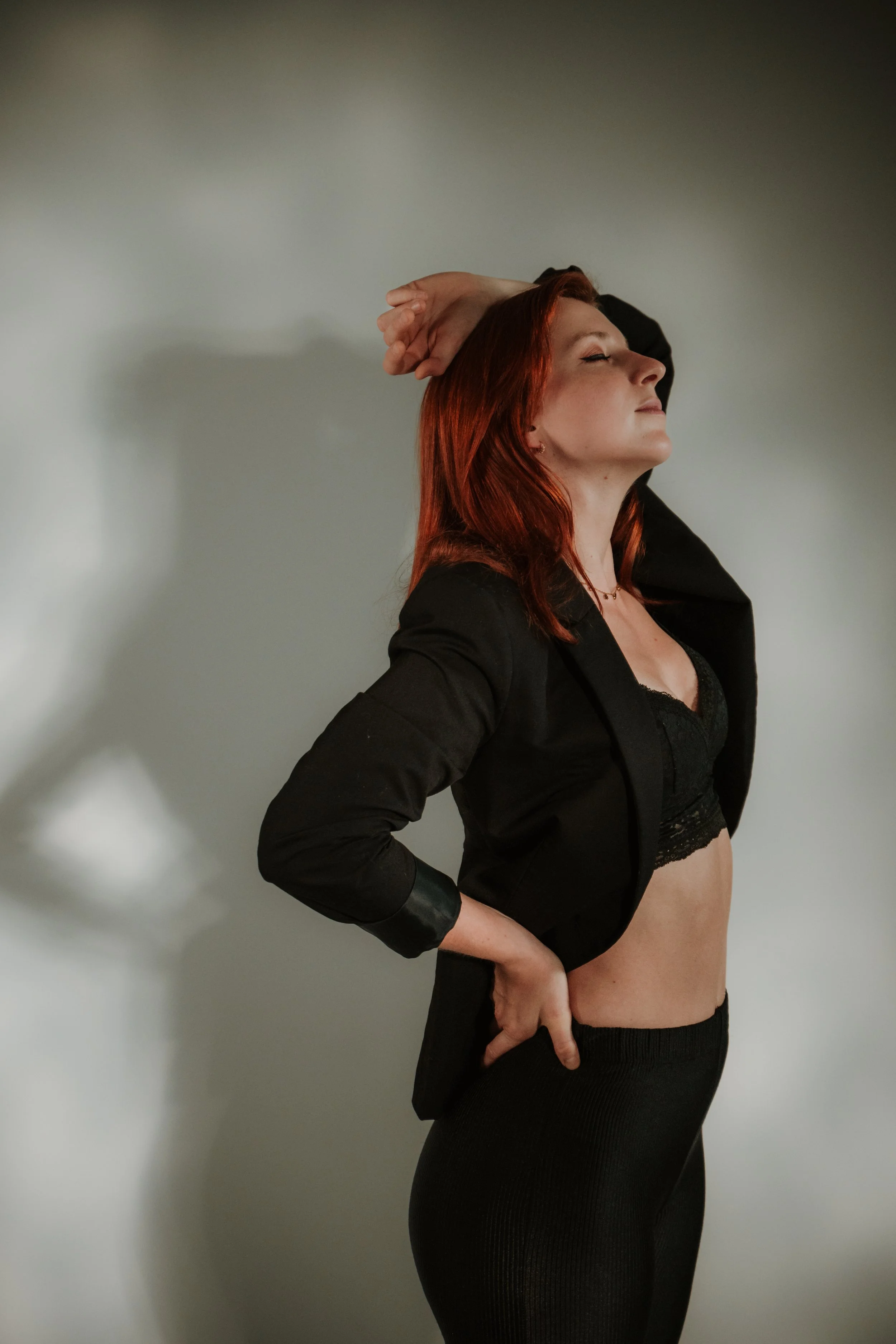 A woman with long red hair and light skin stands in profile with her eyes closed, wearing a black blazer, lace top, and black pants, posing against a plain light background.