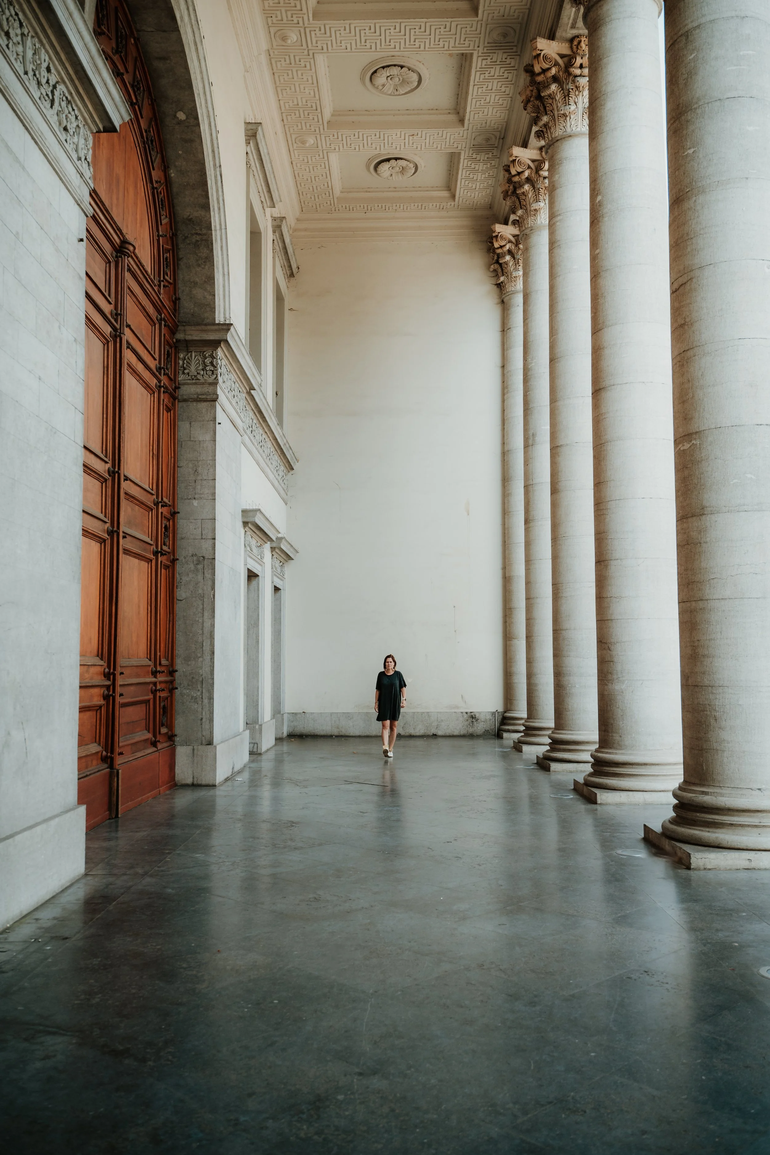 A woman in a black dress walking through a grand architectural corridor with tall columns and a high, decorated ceiling.