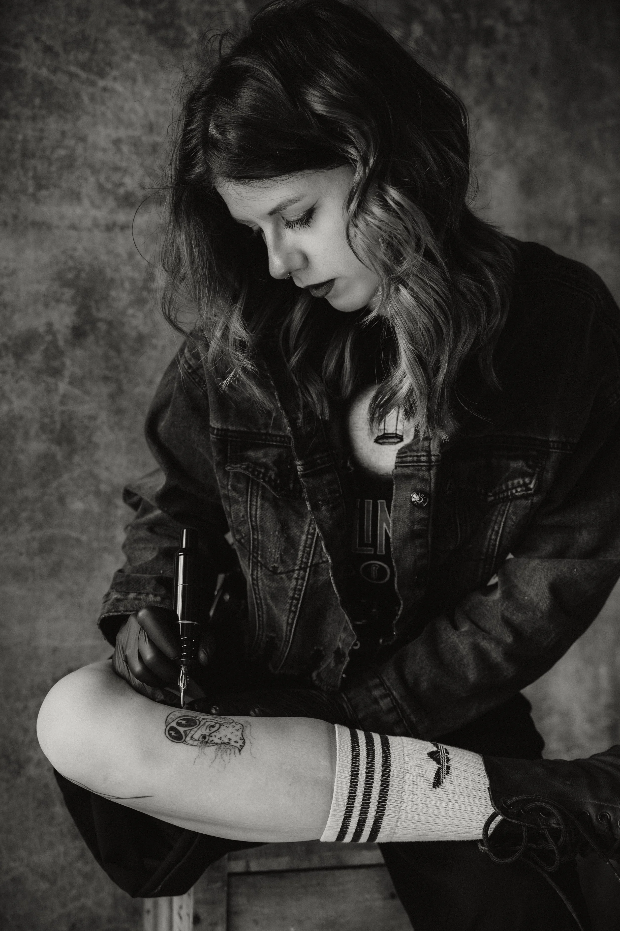 A young woman with dark, wavy hair and dark lipstick, wearing a denim jacket and a graphic t-shirt, is sitting and drawing on her leg with a marker. She has a tattoo on her calf and is wearing striped knee-high socks and black shoes. The background a