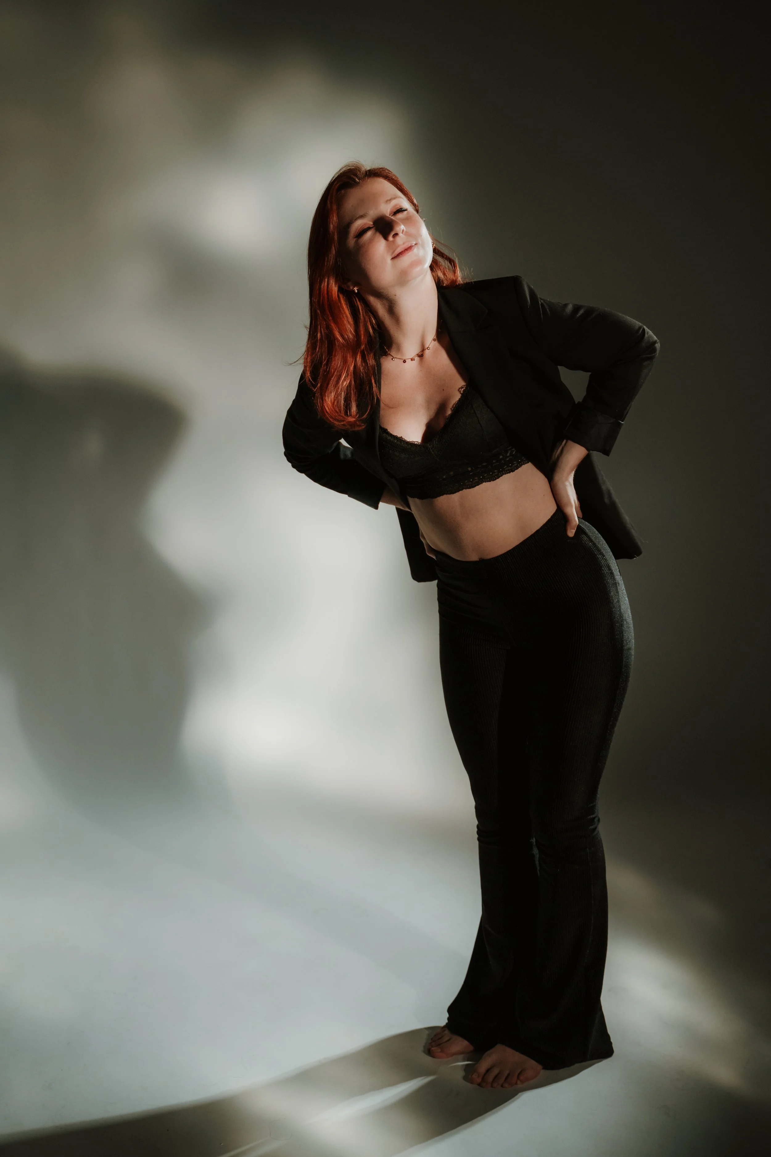 A woman with red hair standing against a dark and light gradient background, wearing black pants, a black top, and an unbuttoned black blazer. She is barefoot, smiling softly with her eyes closed, and has her hands on her hips.