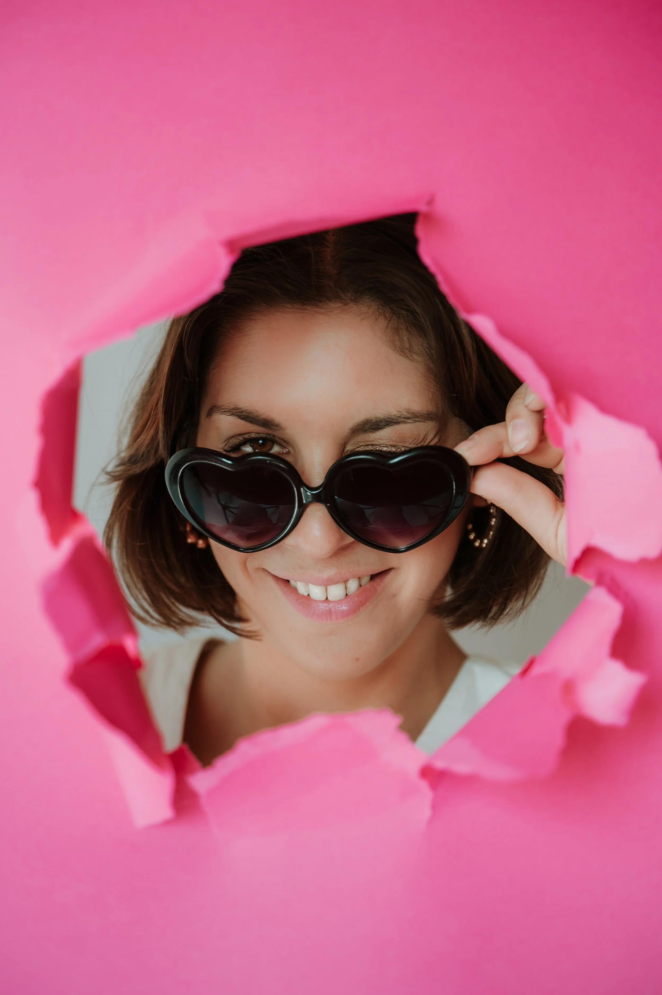 A woman is looking through a torn piece of pink paper, wearing heart-shaped black sunglasses and smiling.