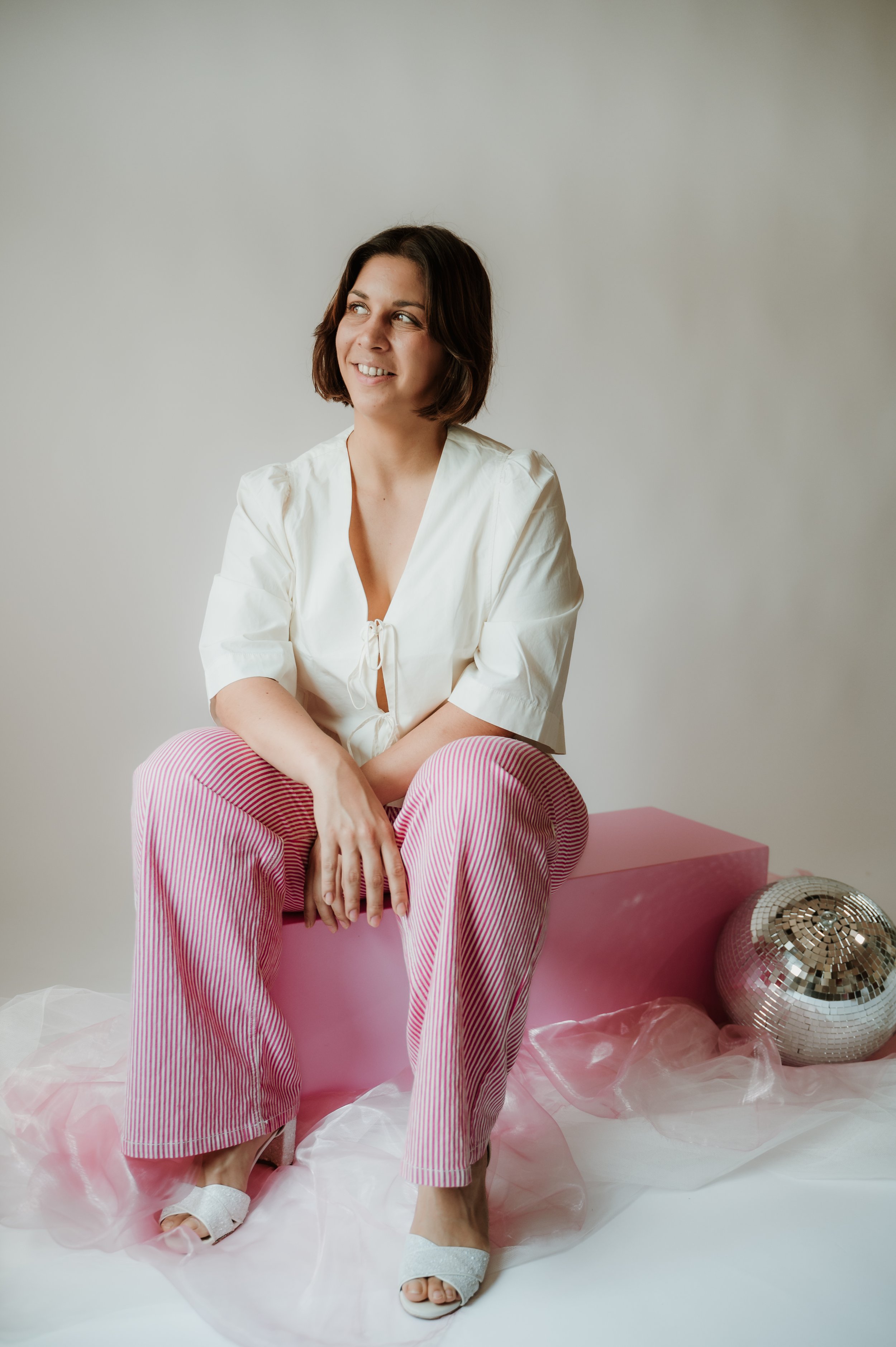 A young woman with shoulder-length brown hair sitting on a pink box, wearing a white blouse, pink and white striped pants, white sandals, smiling and looking to the side. She is surrounded by pink and silver balloons, pink tulle fabric, and a plain w