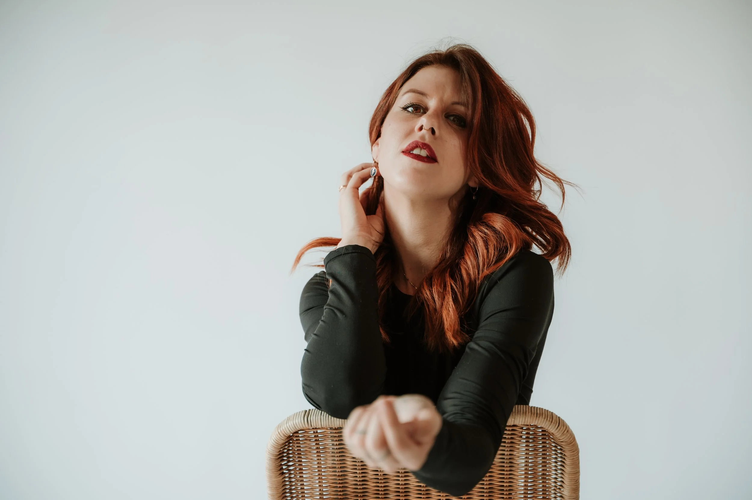 A woman with red hair in a loose wave hairstyle, wearing makeup with dark lipstick and eye makeup, dressed in a black long-sleeve top, sitting on a wicker chair against a plain light gray background, looking at the camera with a confident expression,